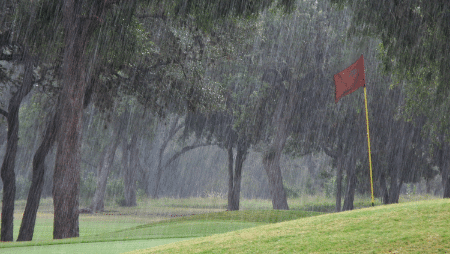 A golf course during a rainstorm with a flag on the green, surrounded by trees and visible rain.