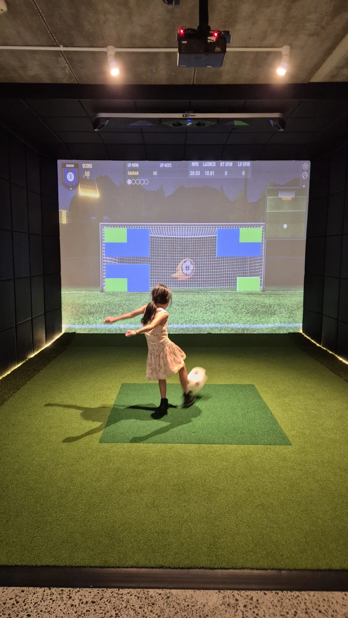 A girl in a beige dress playing soccer on an indoor simulation screen that shows a virtual soccer goal.