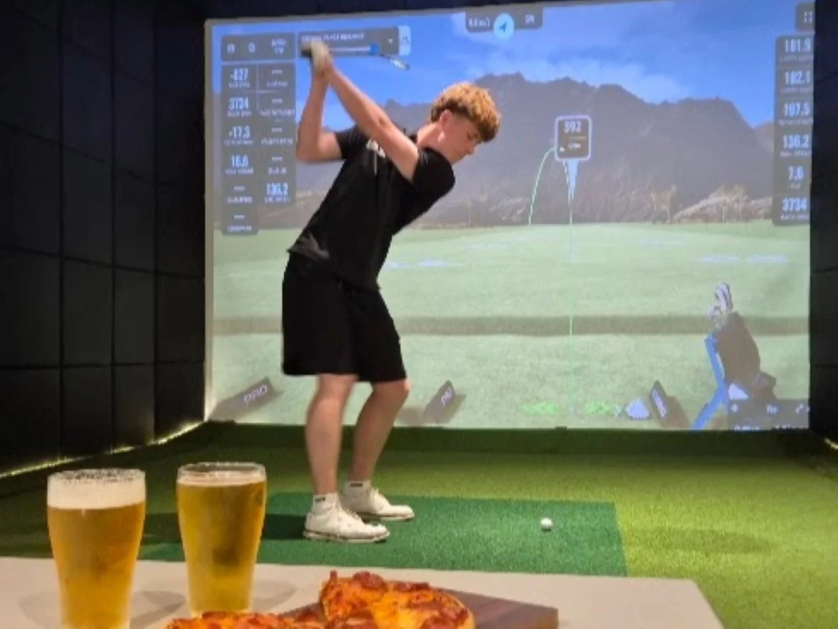 Young man playing indoor golf on a driving range,  two glasses of beer and a pizza on a table in the foreground.