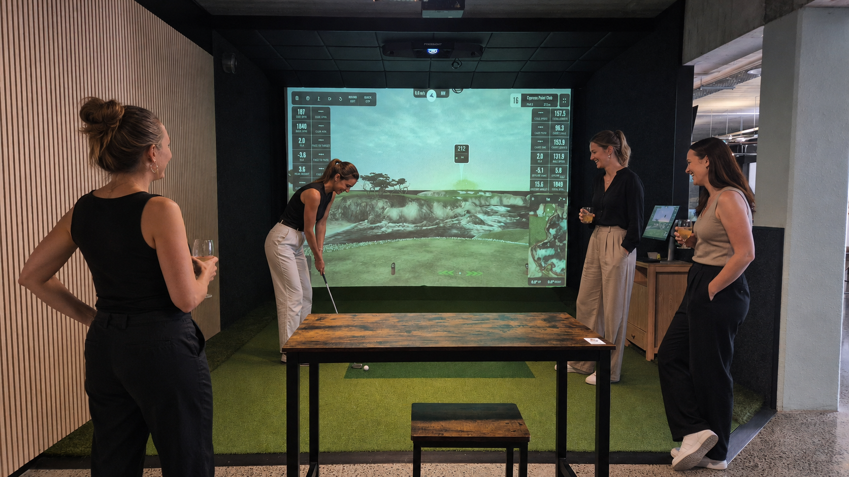 Four women gathered in an indoor golf simulation room. One woman is taking a swing while the other three watch and hold drinks. A large screen displays a virtual golf course.