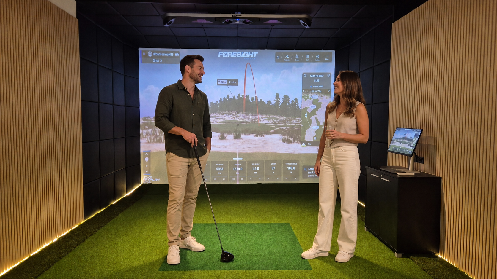 Two people, a man and a woman, in a golf simulation room. The man holds a golf club and is preparing to hit a golf ball, while the woman holds a drink and watches him. There's a large screen displaying a golf course and data about the shot, with a small computer monitor on a black cabinet beside them. The room has a virtual golf environment with wooden and black padded walls, green artificial turf, and LED lighting.