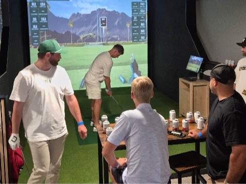People at a golf simulator facility, with one person swinging a golf club on a screen and others watching.