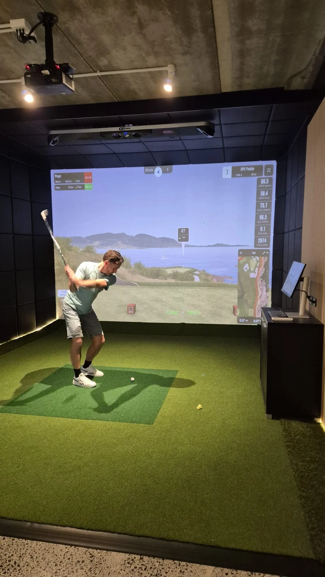Person practicing golf swings indoors on a simulator with a large screen showing a golf course landscape.