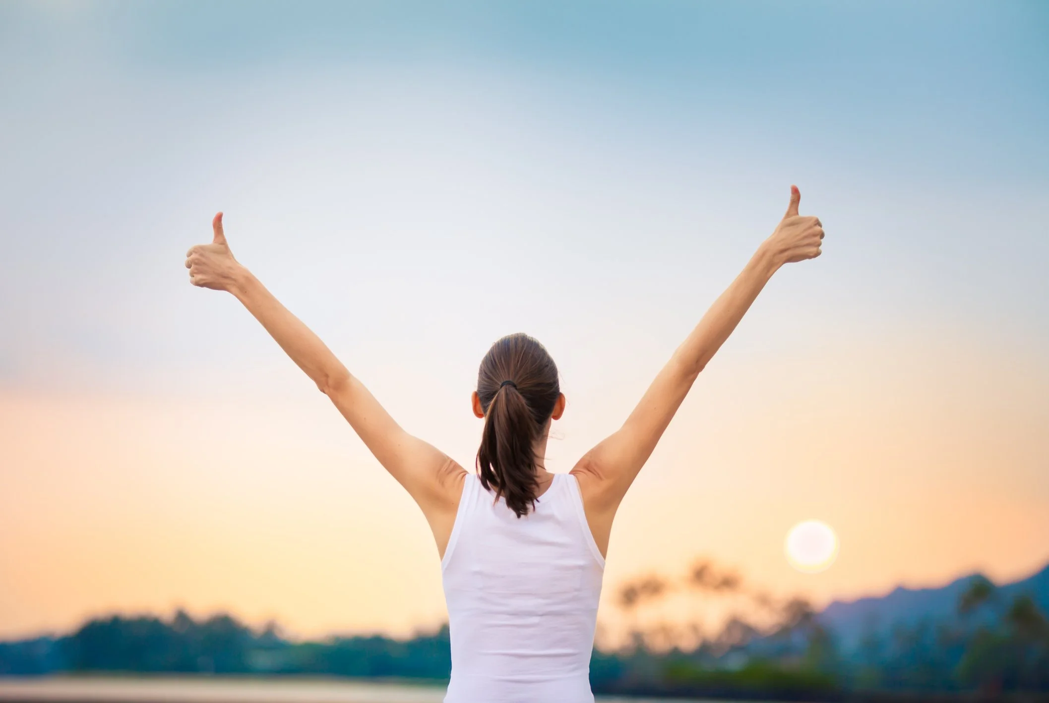 A woman with brown hair tied in a ponytail raising her arms in a 'V' shape with thumbs up gestures against a sunset sky, over a rural landscape.