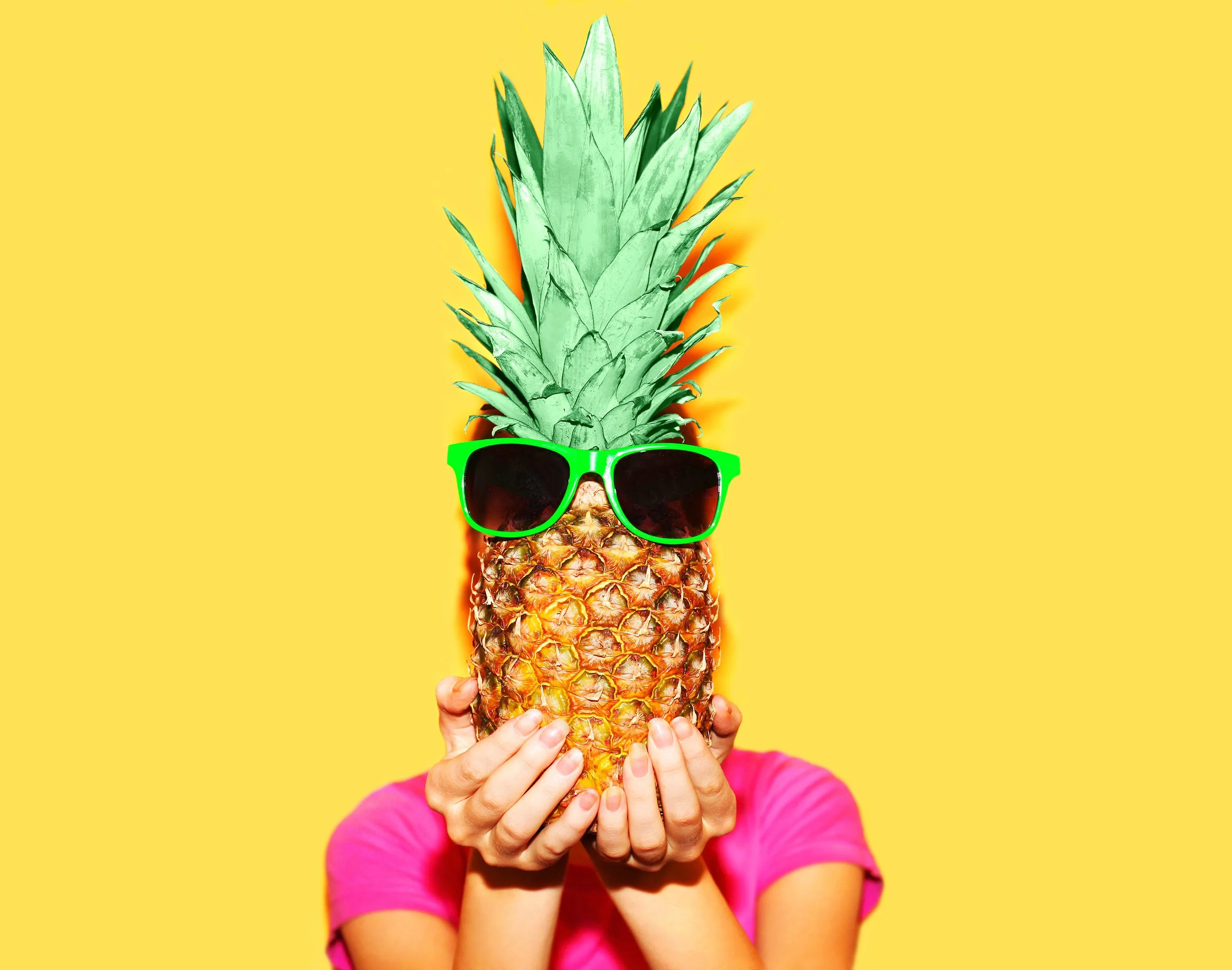 A person holding a pineapple wearing green sunglasses and a pink shirt against a yellow background.
