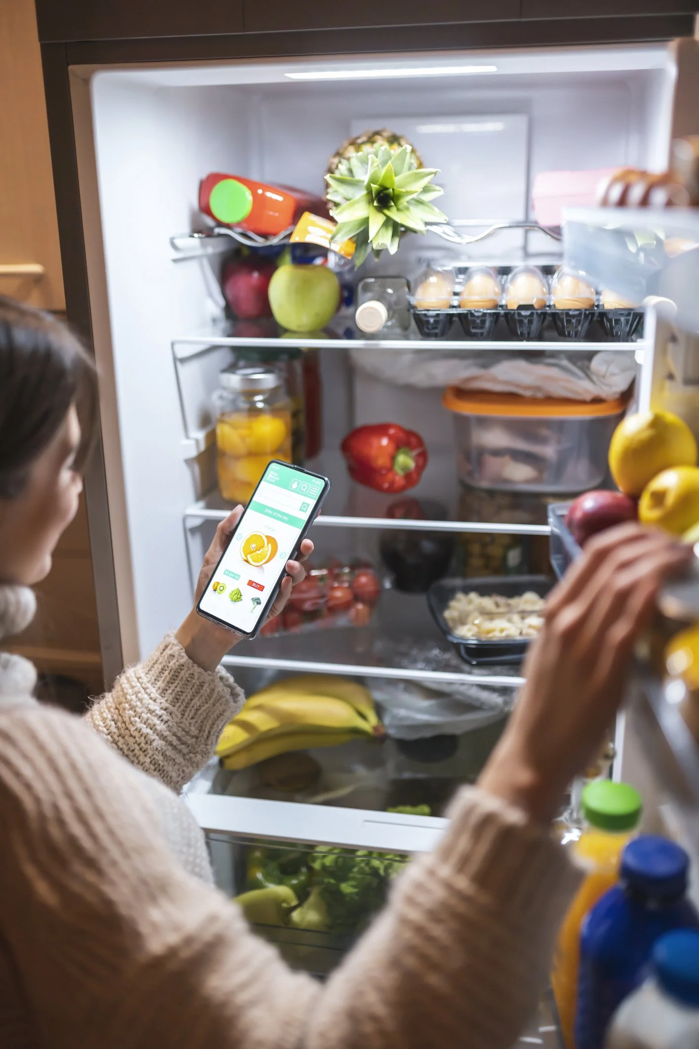 Person in a beige sweater using a smartphone to view a grocery shopping app while standing in front of an open refrigerator filled with fruits, eggs, vegetables, and other food items.