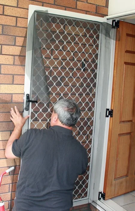 Man inspecting a white security screen door with decorative metal mesh beside an open wooden door.
