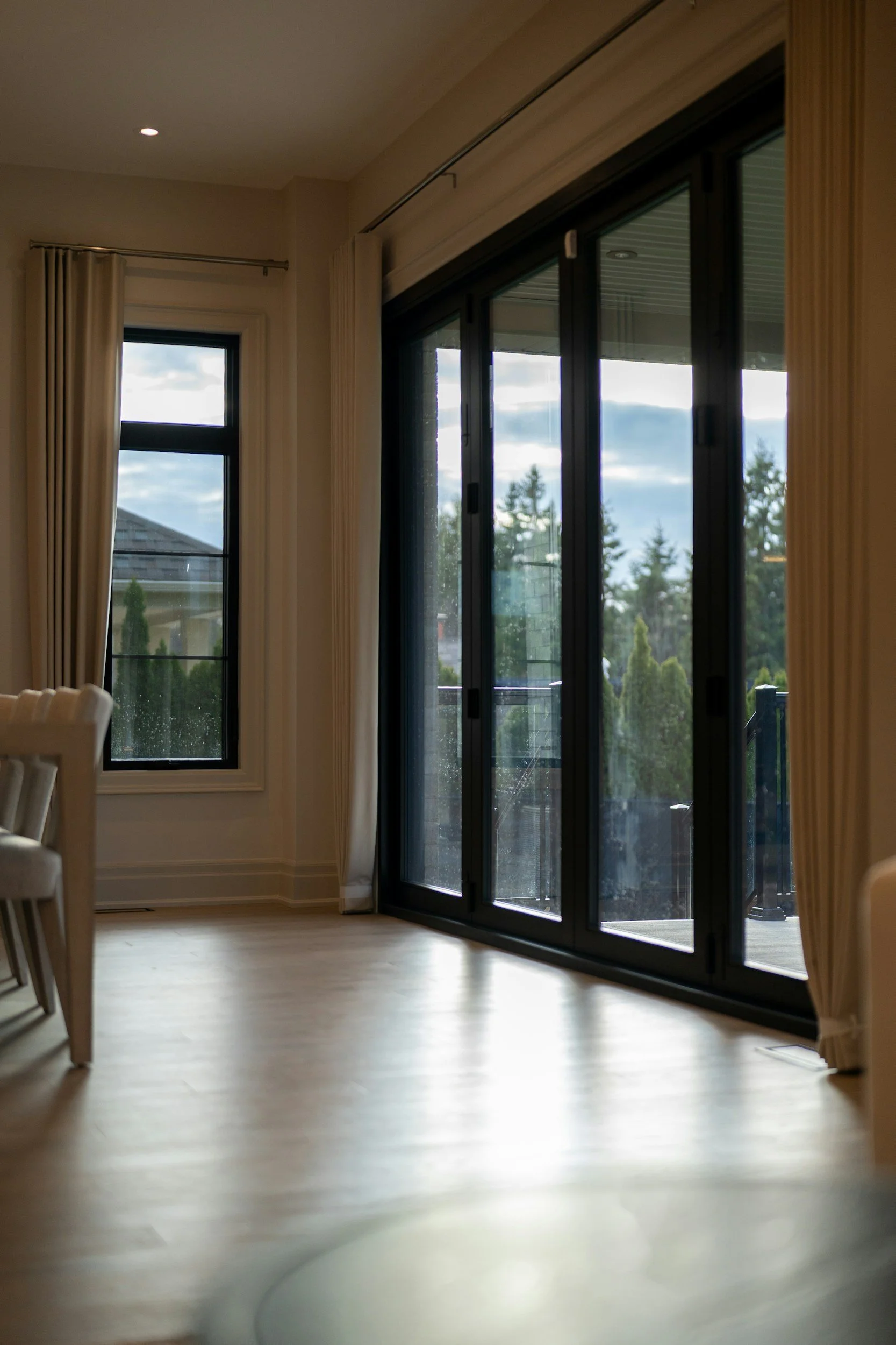 Bright dining room with floor-to-ceiling black-framed glass doors, a curtained window, and outdoor greenery visible beyond.