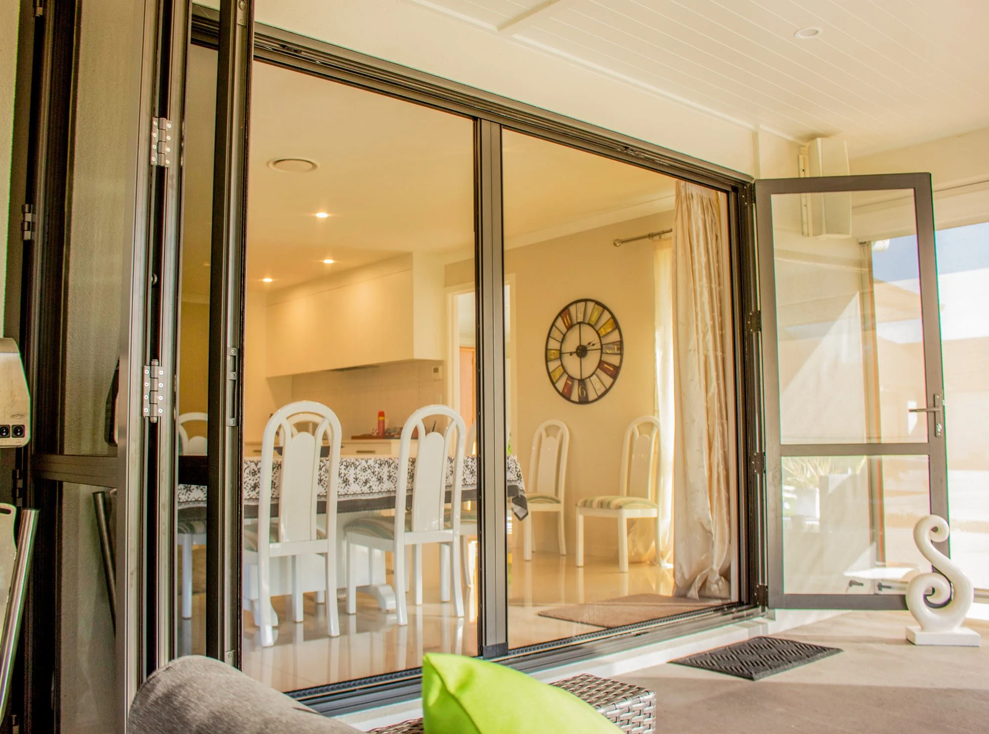 Bright home interior with folding glass doors, white dining chairs, wall clock, curtains, and patio seating