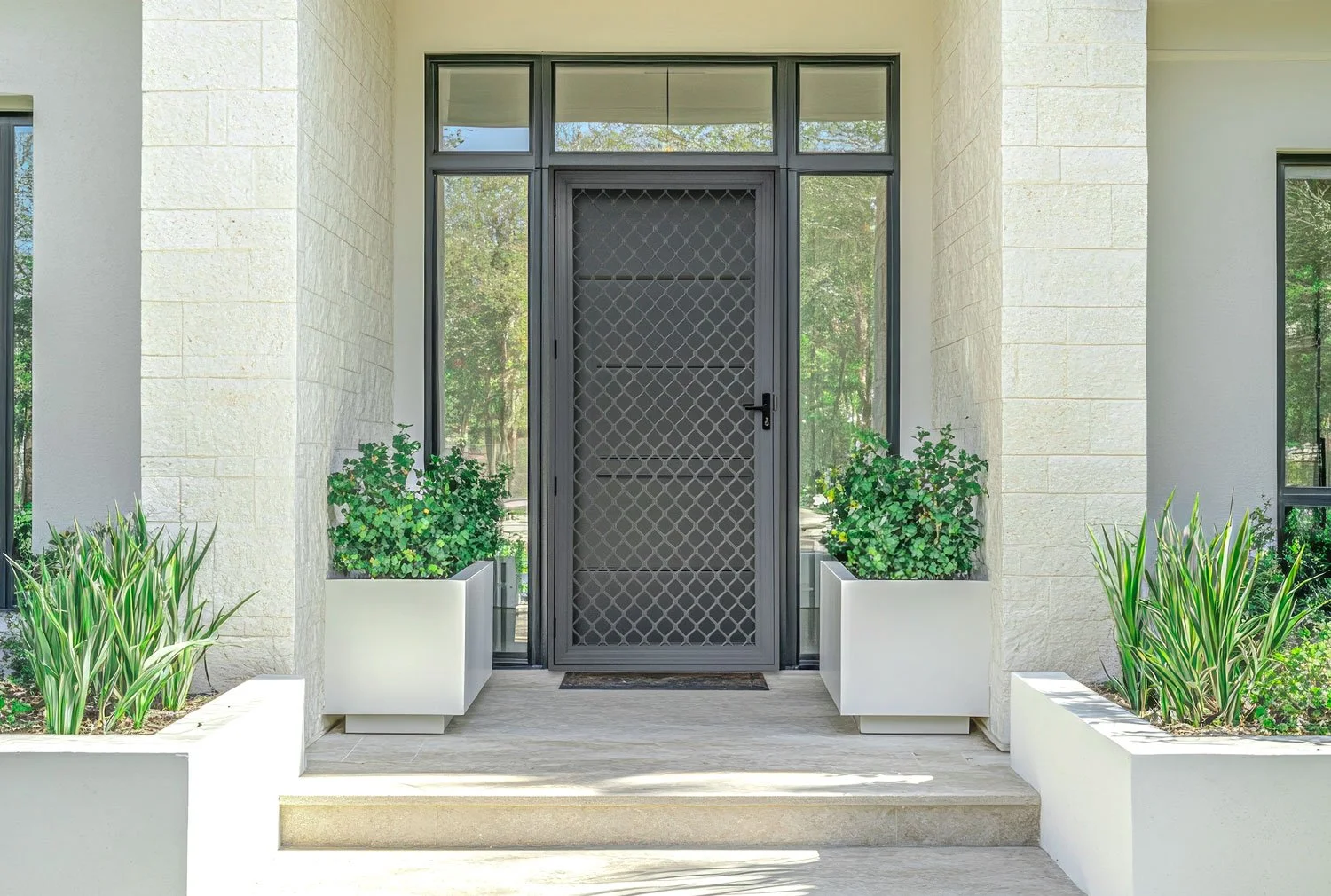 Modern home entrance with a gray security door, surrounding windows, white stone walls, and potted green plants