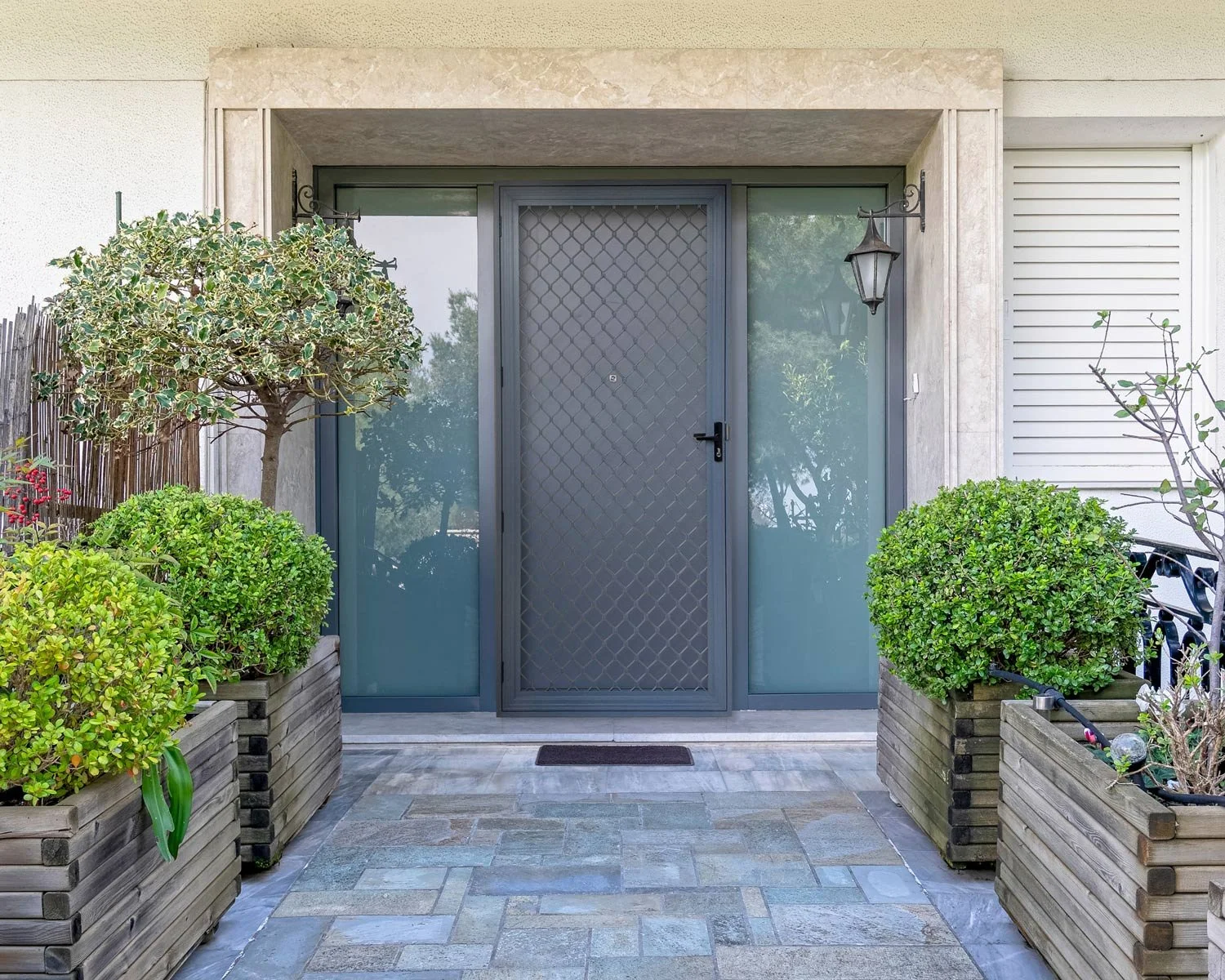 Modern home entryway with gray security door, sidelights, wall lanterns, and landscaped planter boxes