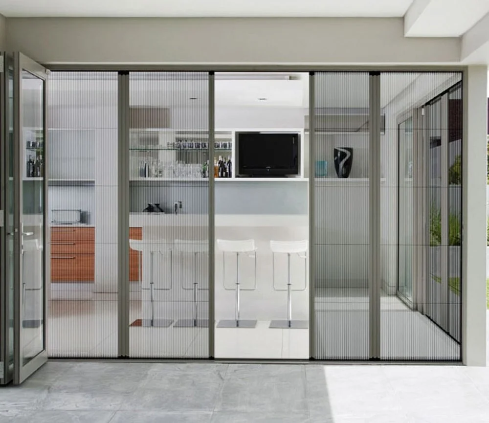 Modern open-plan kitchen and living area visible through floor-to-ceiling glass doors, with white bar stools, built-in television, and display shelving.