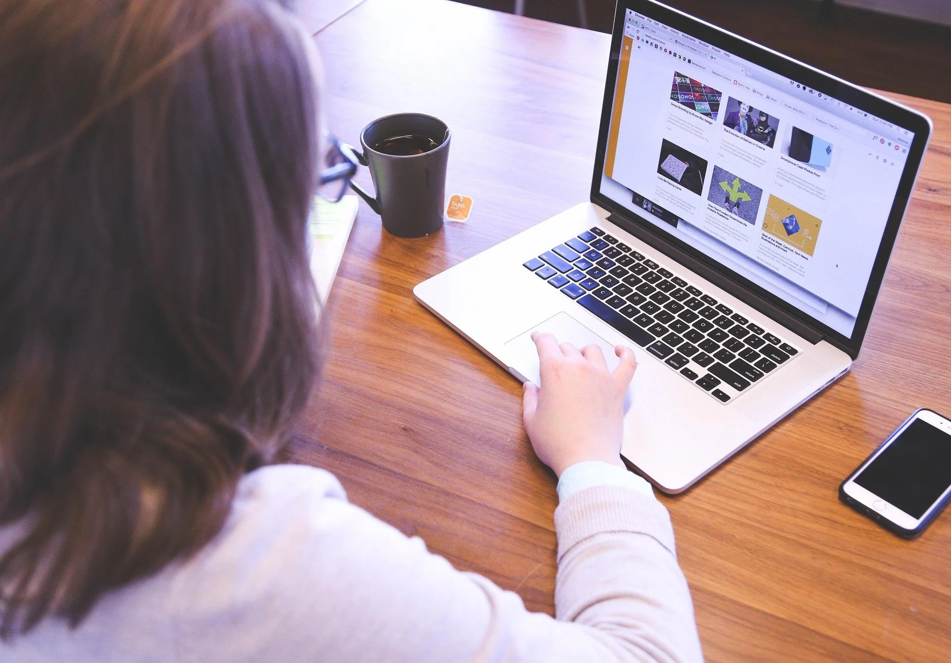 A person with long brown hair wearing glasses is working on a silver laptop on a wooden desk. The laptop screen displays a webpage with articles and images. There is a black coffee mug with a tea bag label hanging on the side, a smartphone next to the laptop, and some notebooks on the desk.