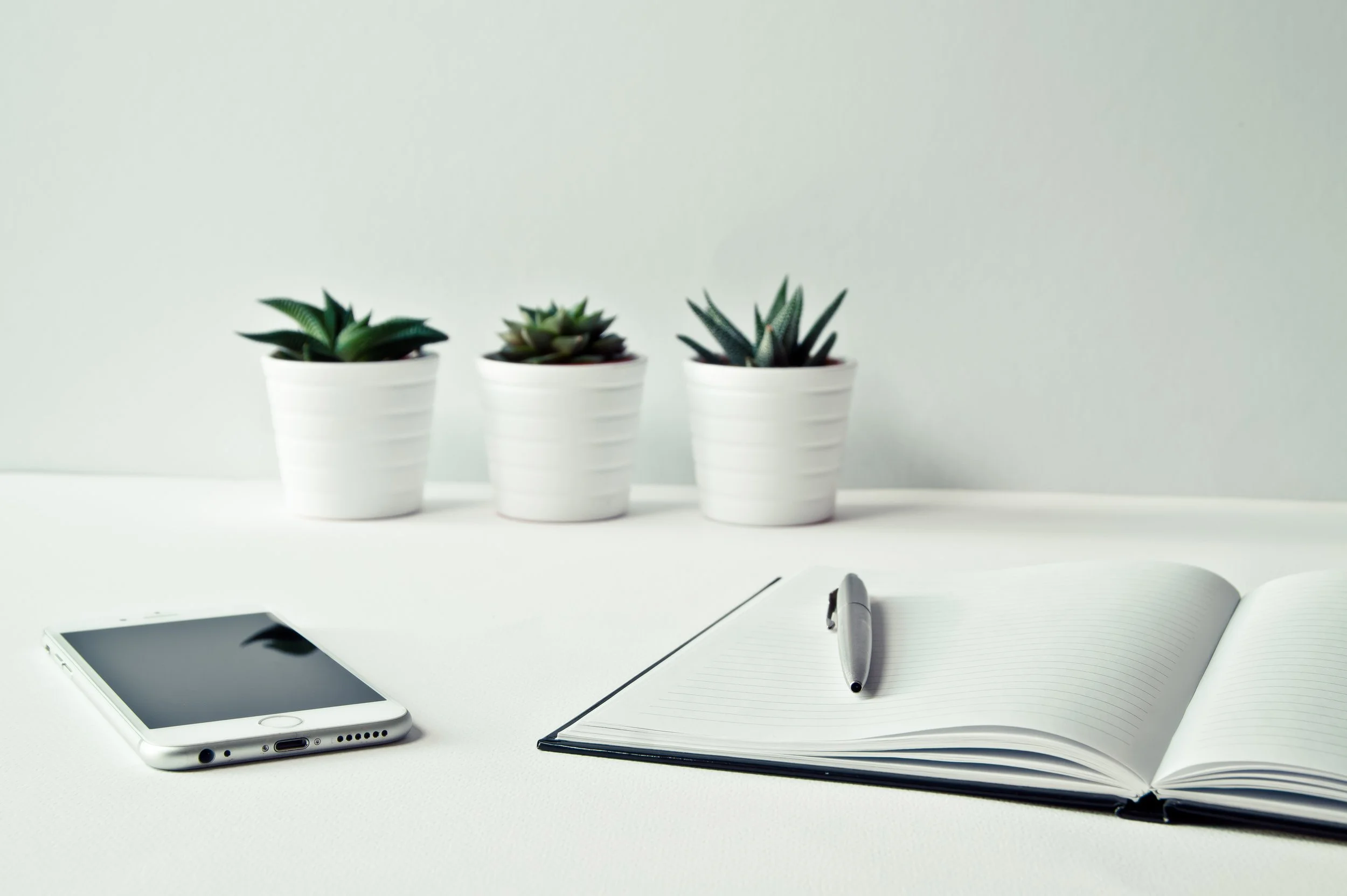 Three potted succulents on a white desk, an open notebook with a pen, a smartphone, and a notebook in the foreground.