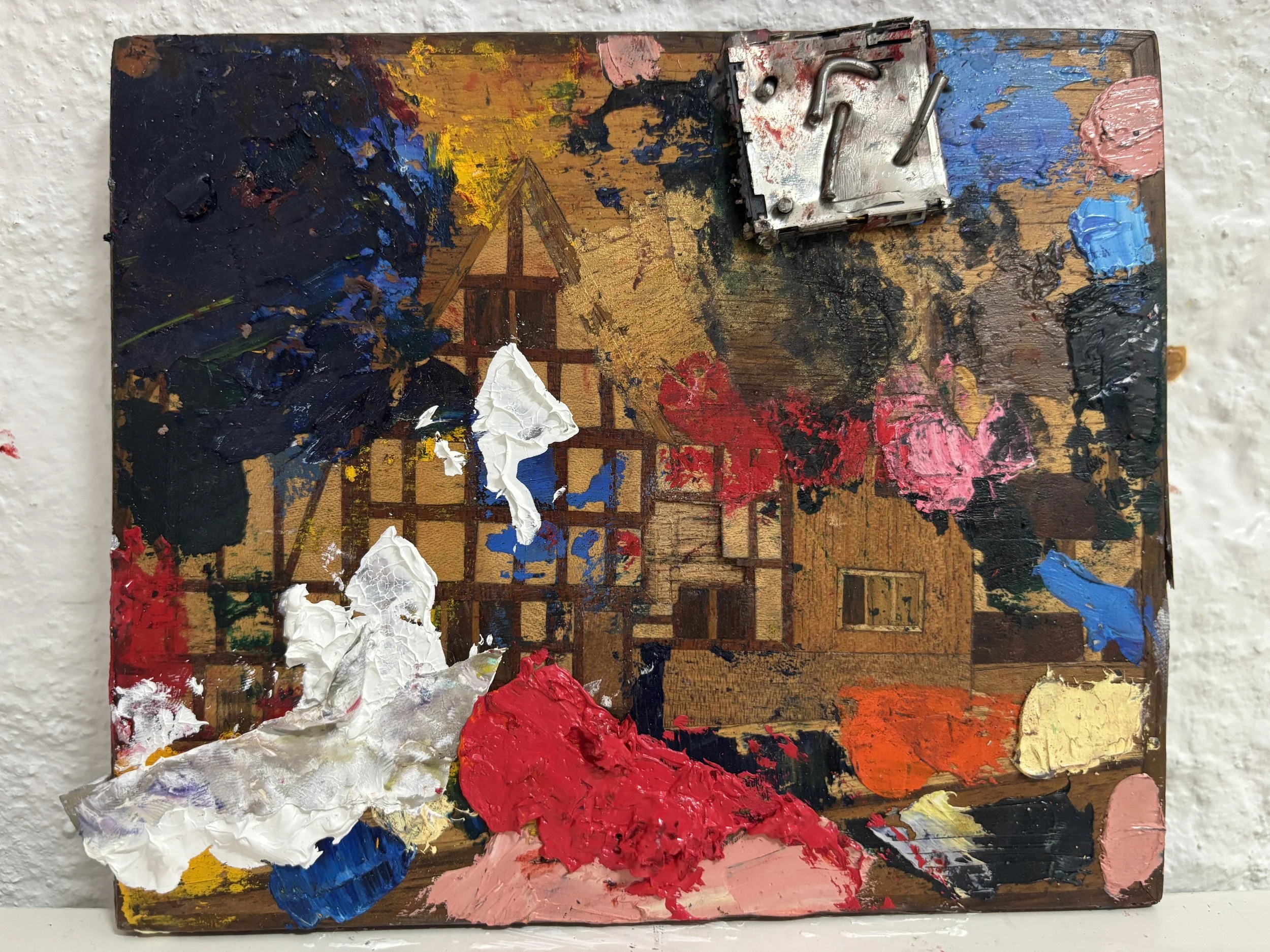 A small, abstract, colorful painting on a wooden surface, with thick application of paint in various colors including white, red, blue, pink, yellow, and black. There is a small, metallic, mechanical object attached to the top right corner of the pai
