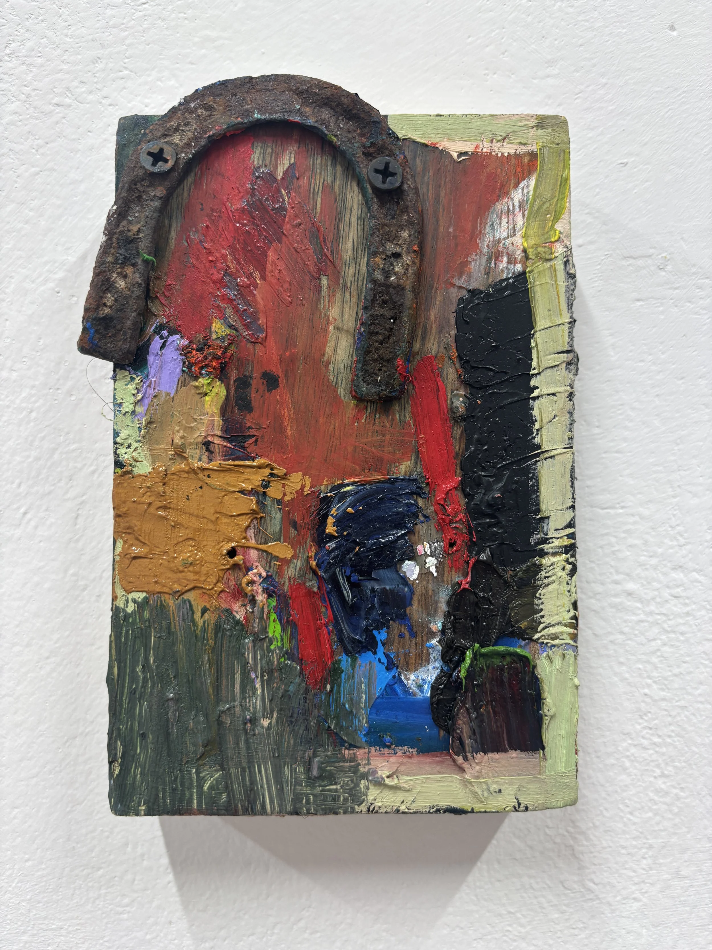 A small, rectangular wooden palette with various colors of thick, textured paint including red, black, yellow, green, purple, and blue, used in abstract or mixed media art, mounted on a white wall.