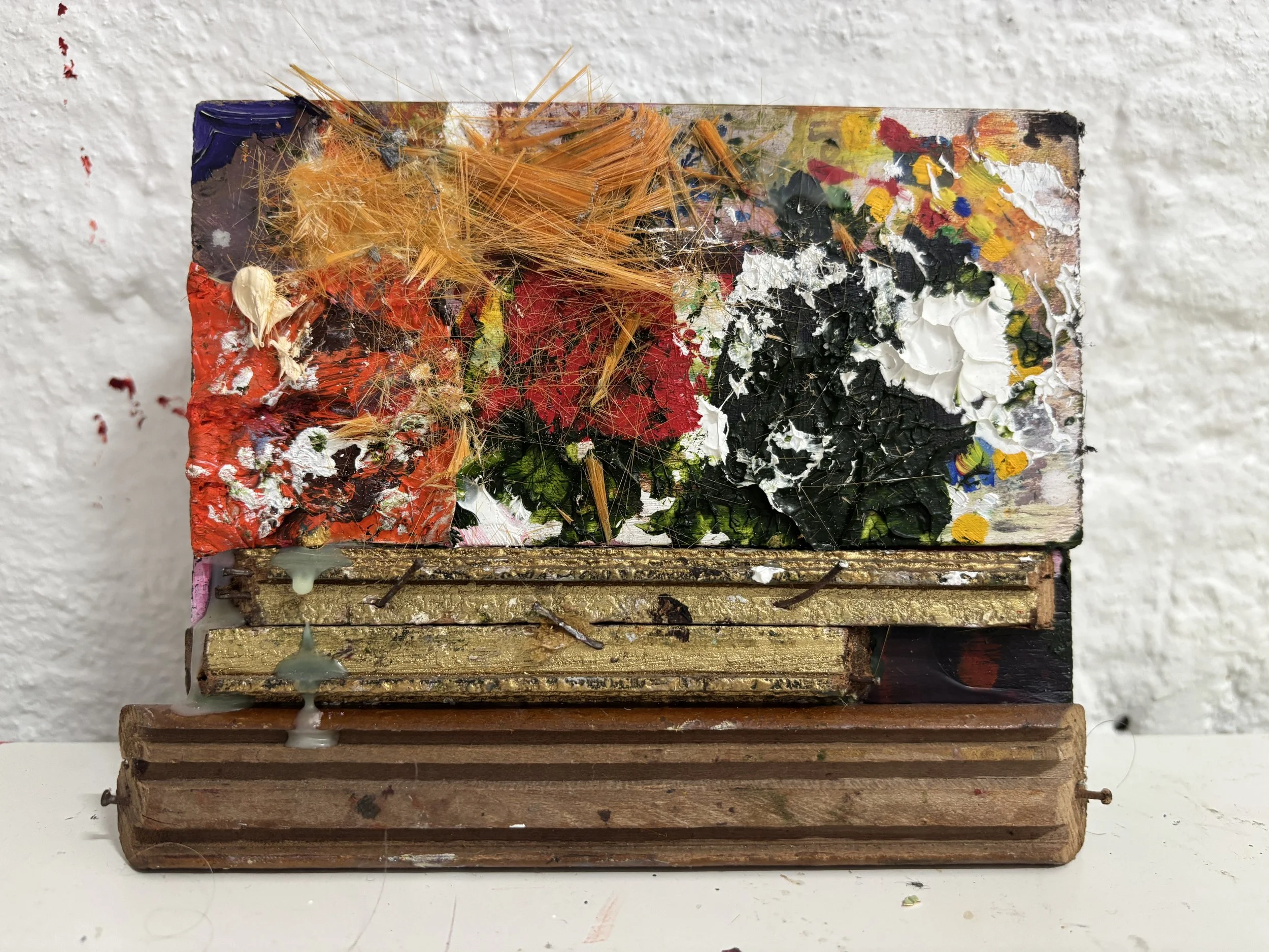 A wooden artist's palette with various paint colors and paint smudges, resting on a white surface, against a white textured wall.