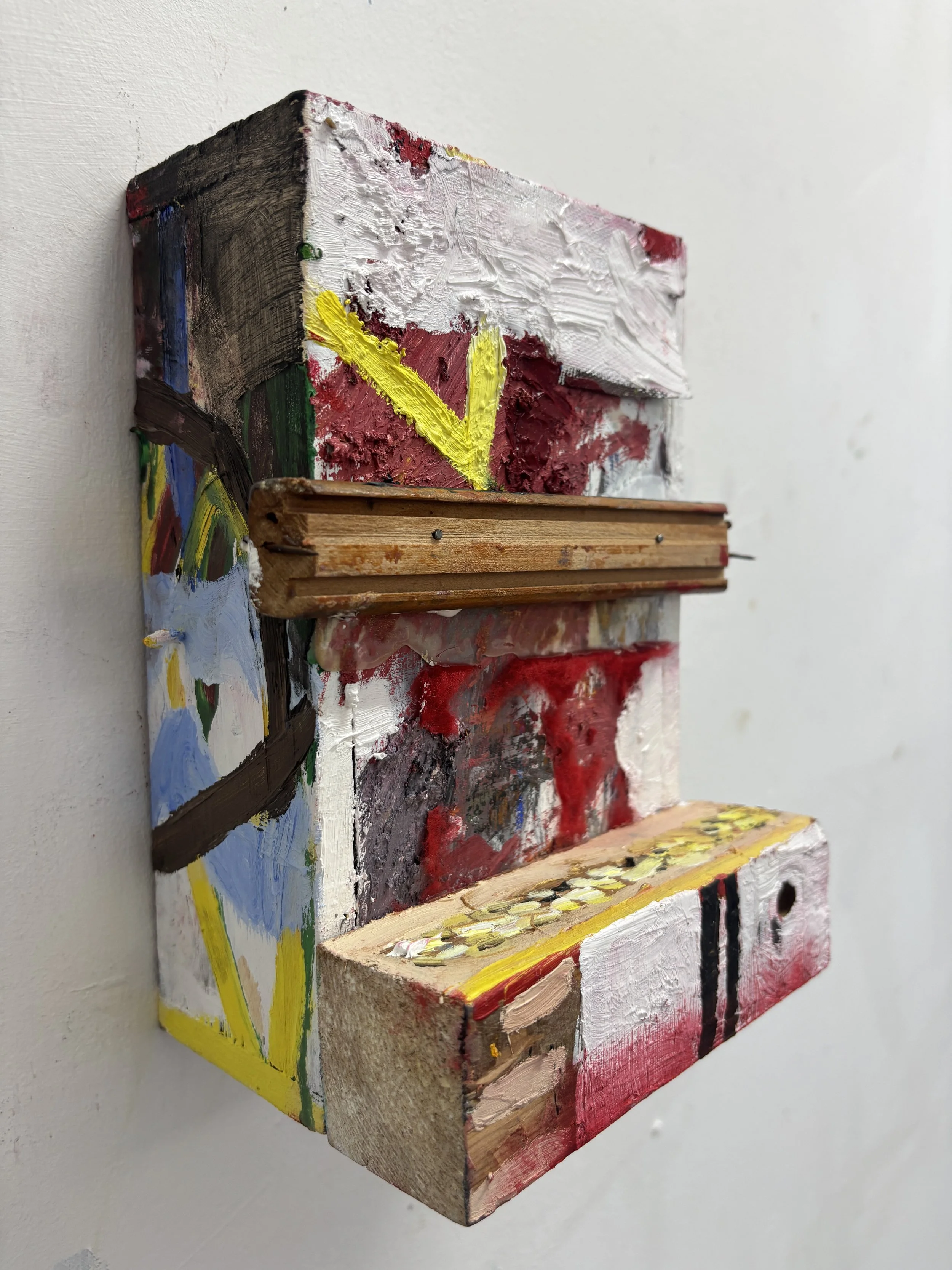 Close-up of a small, colorful, mixed-media abstract painting on a wall, featuring thick layers of paint with visible brushstrokes and textures.