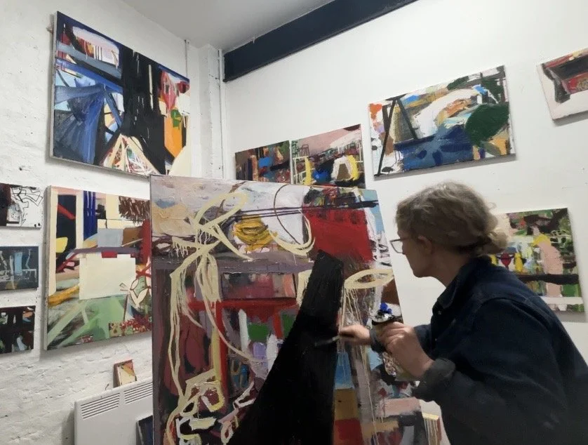 An artist working on a colorful abstract painting in an art gallery with multiple abstract paintings on the white brick walls.