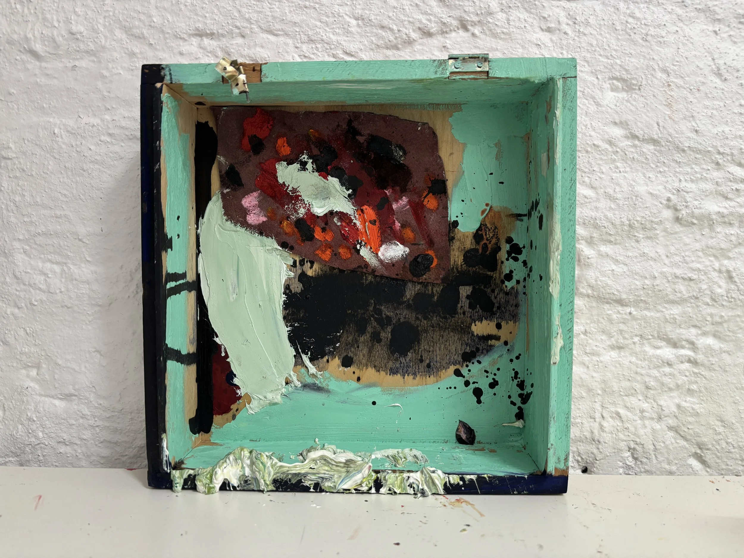 A small, colorful abstract painting on a simple wooden box with a teal border, decorated with splatters and patches of various paint colors, positioned against a white textured wall.