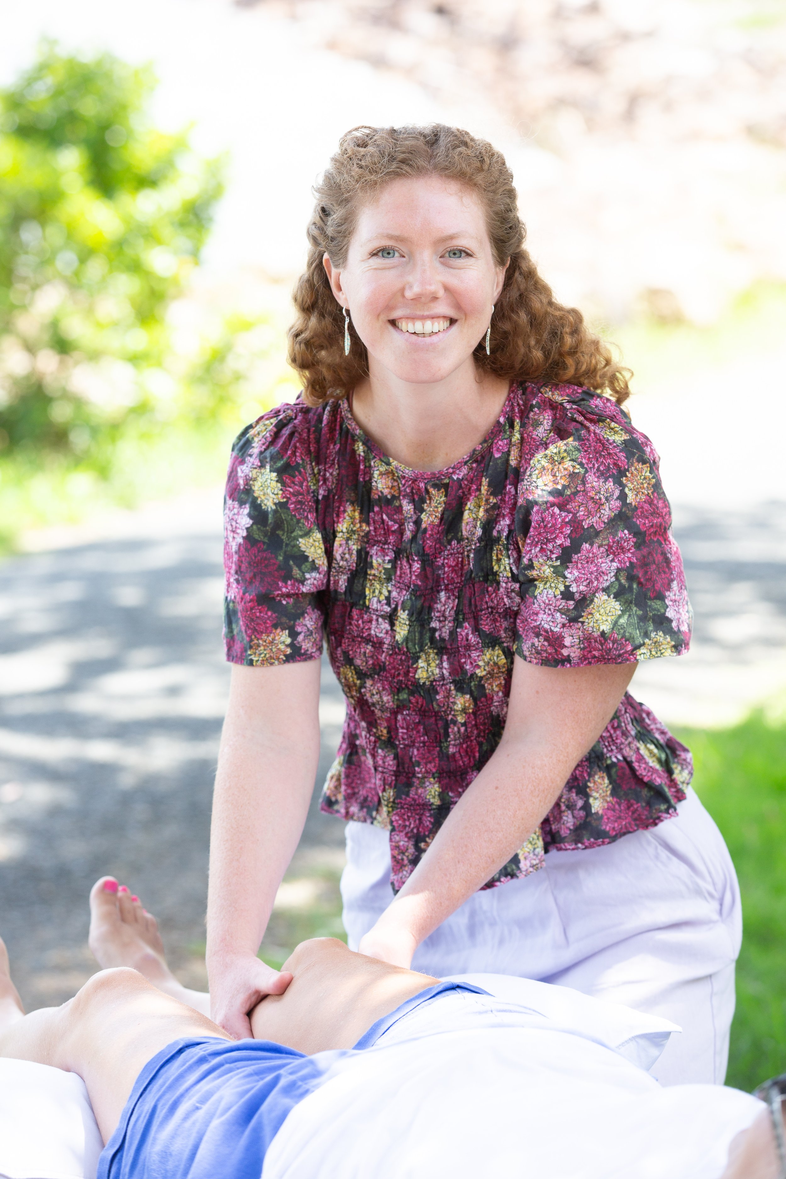 A woman with curly red hair, wearing a floral top and earrings, is outdoors smiling while giving an osteopathic treatment to a person lying on a massage table. The background features a sunny outdoor setting with green foliage and a pathway.