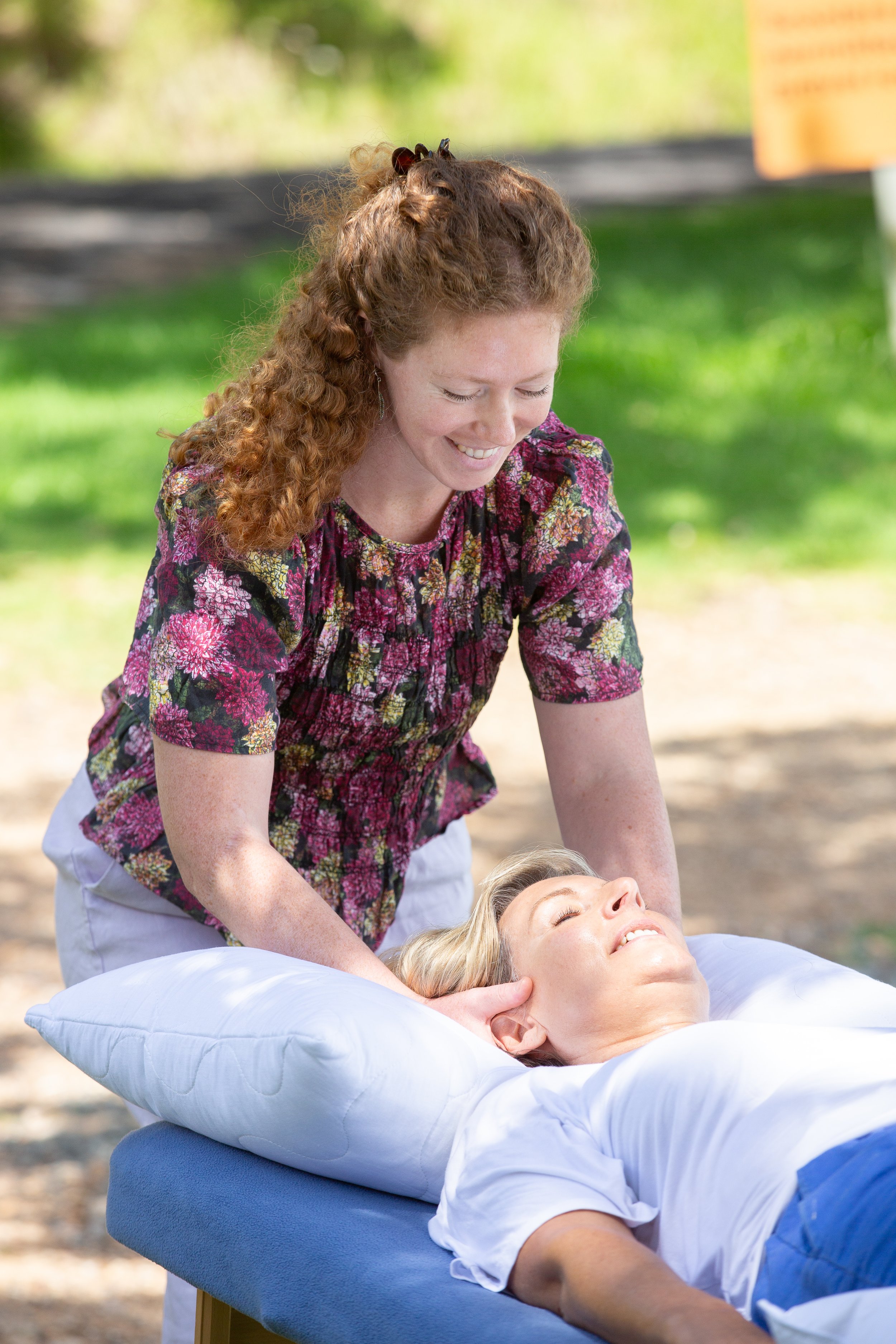 A woman giving an osteopathic treatment outdoors to a woman lying on a massage table, surrounded by greenery.