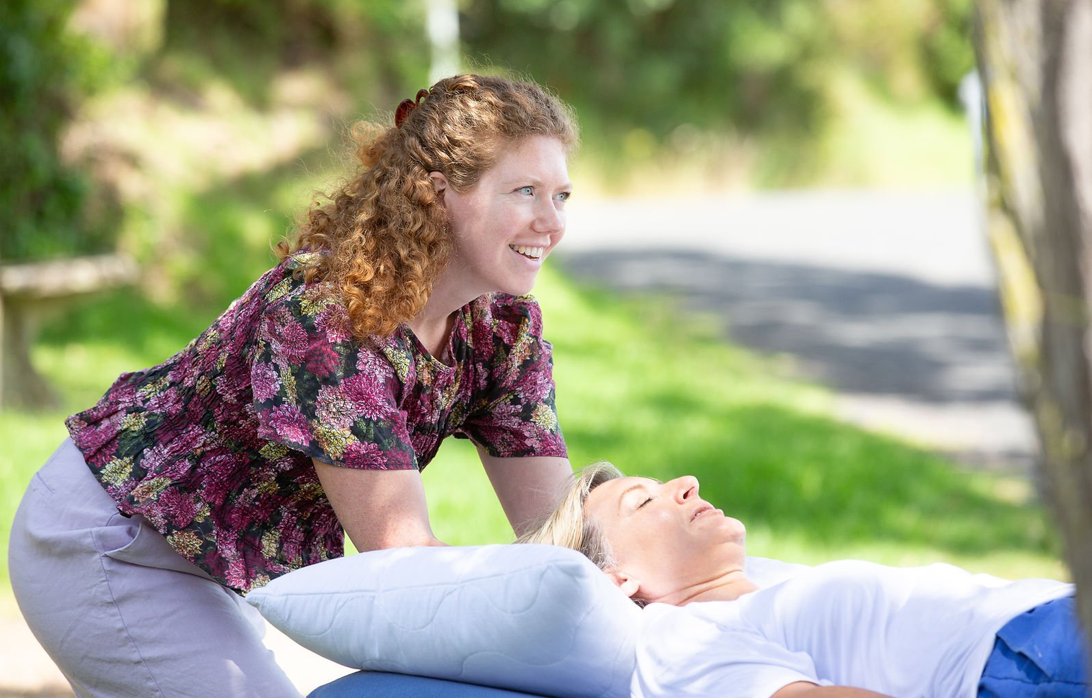 A woman with curly red hair gives a massage to another woman with blonde hair, lying on a table outdoors surrounded by green trees and grass, with a sunny background.