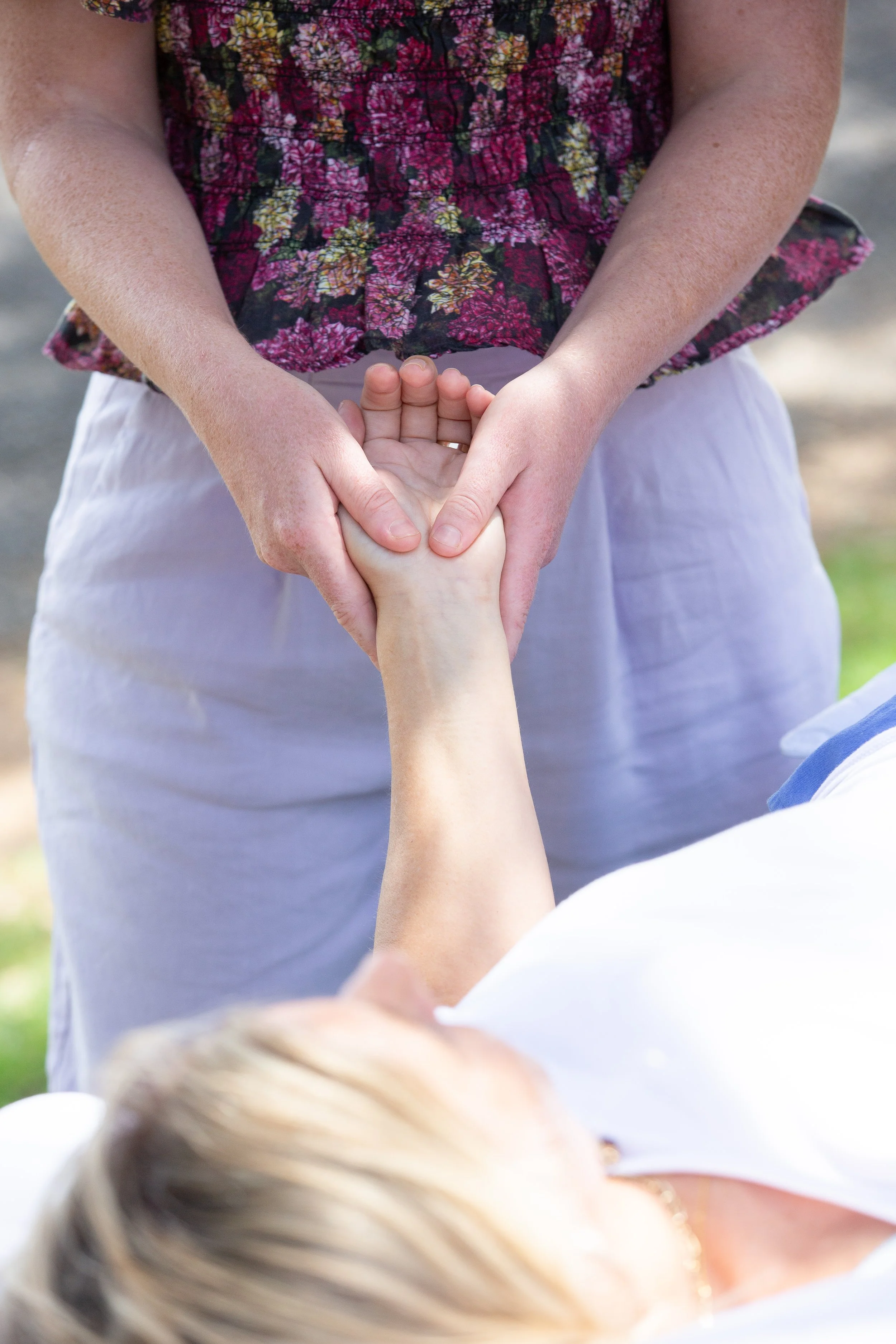 A osteopath in a floral top giving osteopathic treatment to a woman's wrist, both outdoors during daytime.
