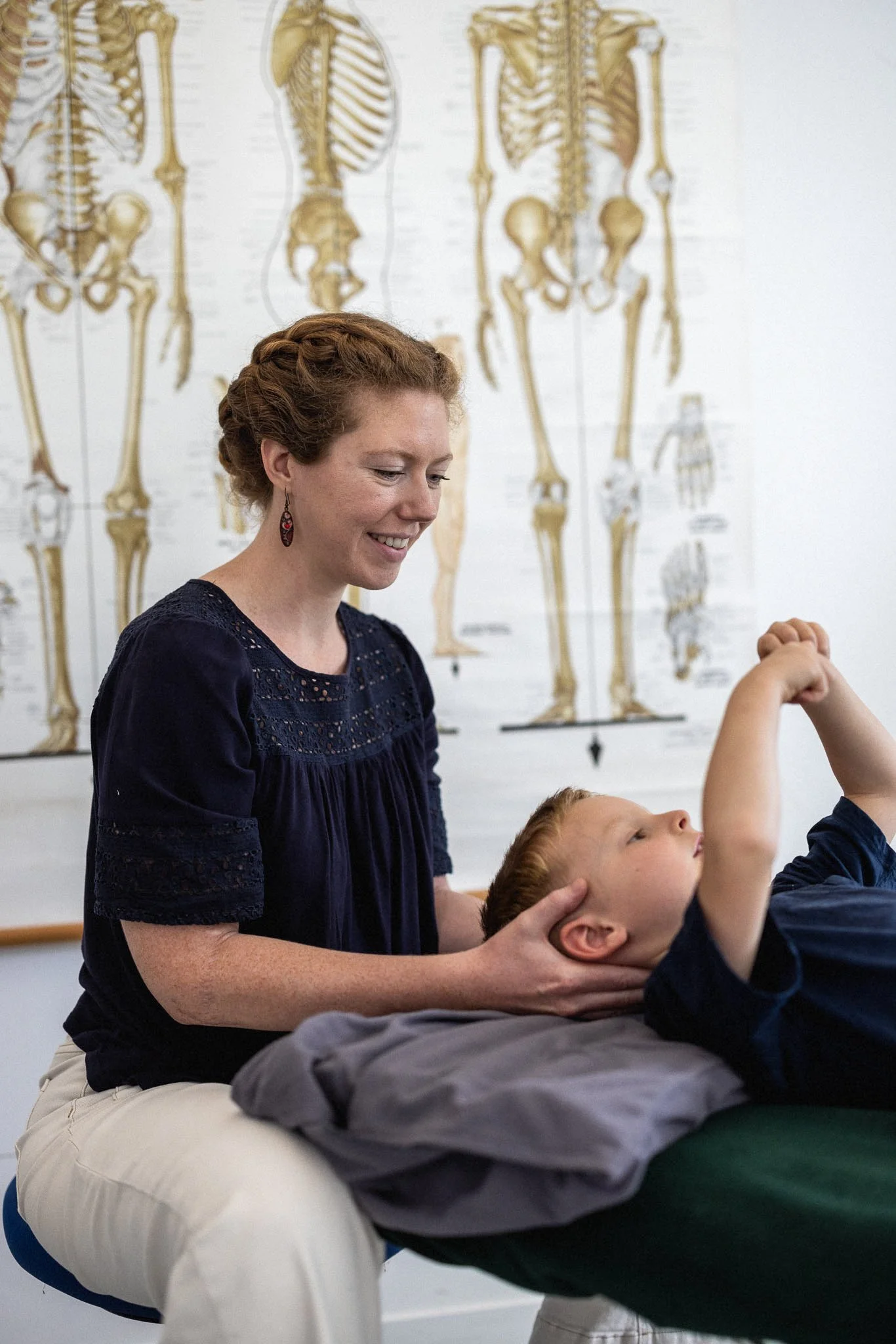 A woman with red hair styled in a braid, smiling, wearing earrings and a navy blue blouse, assisting a young boy with red hair in a medical or physical therapy session, with anatomical posters of the human skeleton on the wall in the background.