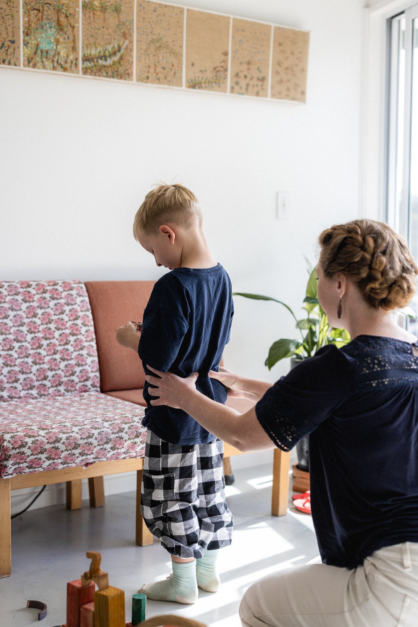 A woman is holding a young boy by the waist as he stands on a rug in a living room, looking at a colorful wooden block toy on the floor. The woman has red hair styled in braids and is wearing a dark blouse and light-colored pants. The boy has short blonde hair, a navy shirt, checkered shorts, and knee-high socks. There are some toys on the floor, a floral-patterned couch, and a framed artwork on the wall.