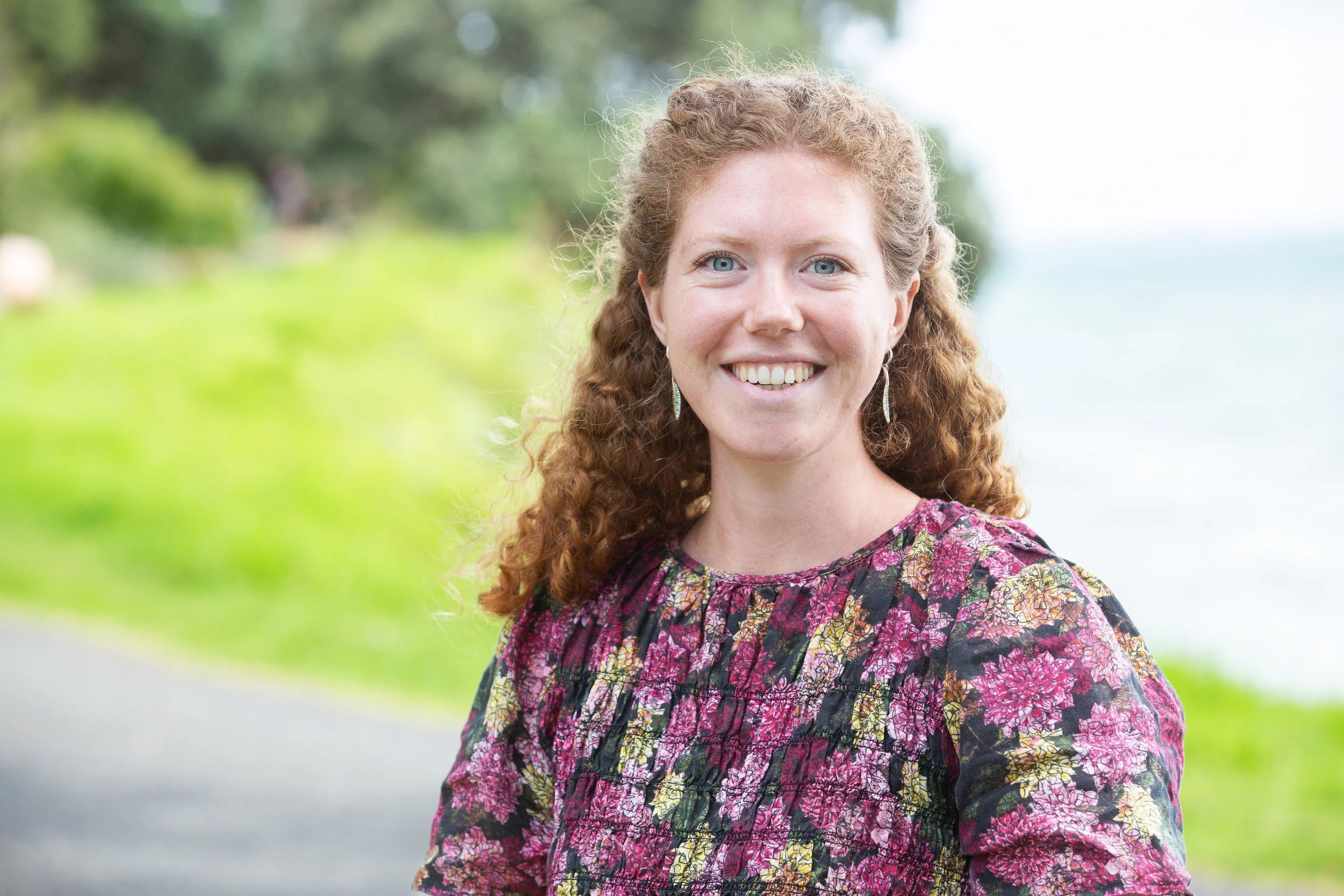 Smiling woman with curly red hair wearing earrings and a multicolored floral dress standing outdoors near a lake or river with greenery in the background.