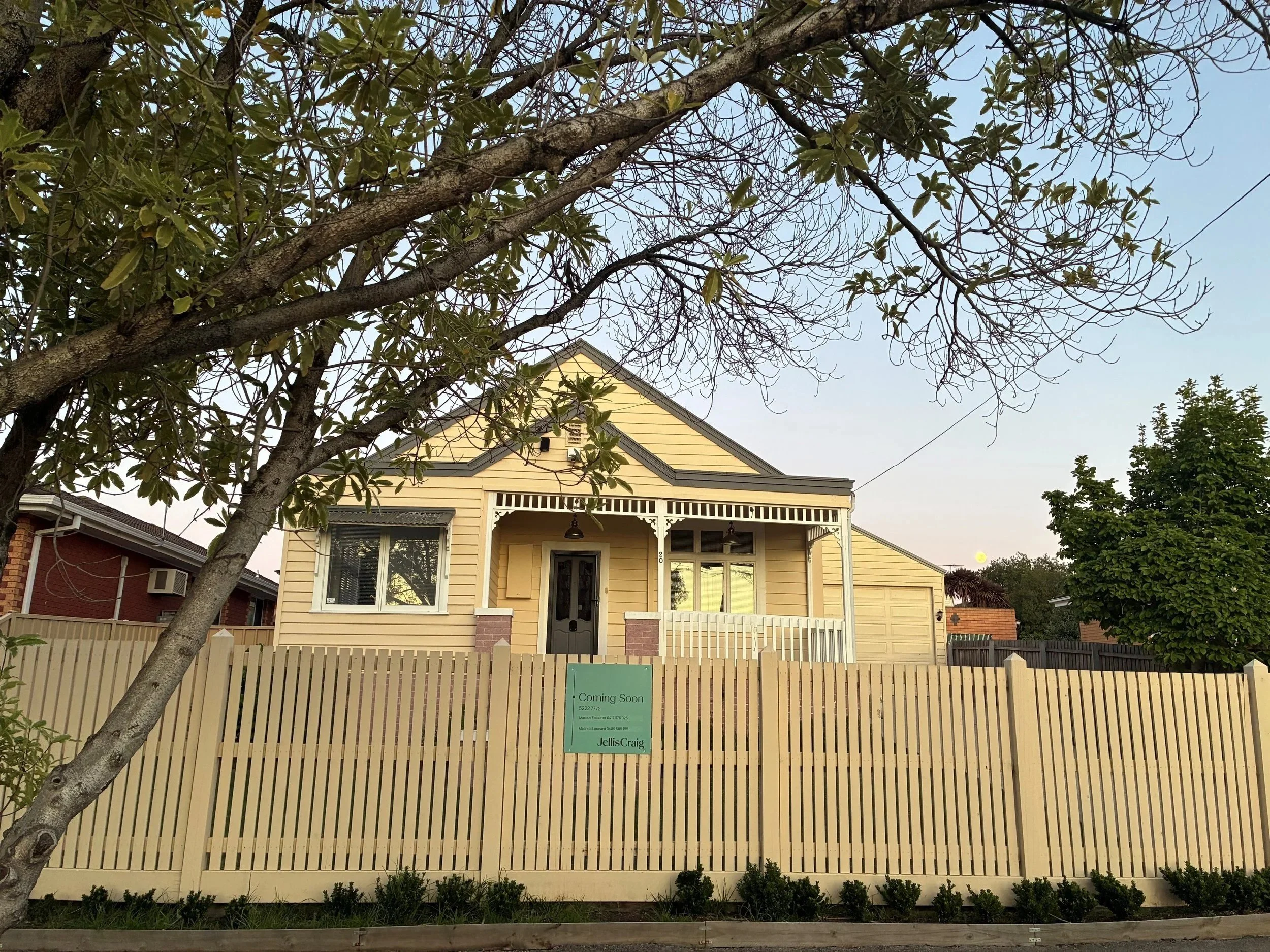 Front view of a yellow house with a front porch, surrounded by a light-colored picket fence, a tree in the foreground, and a 'Coming Soon' sign in the yard.