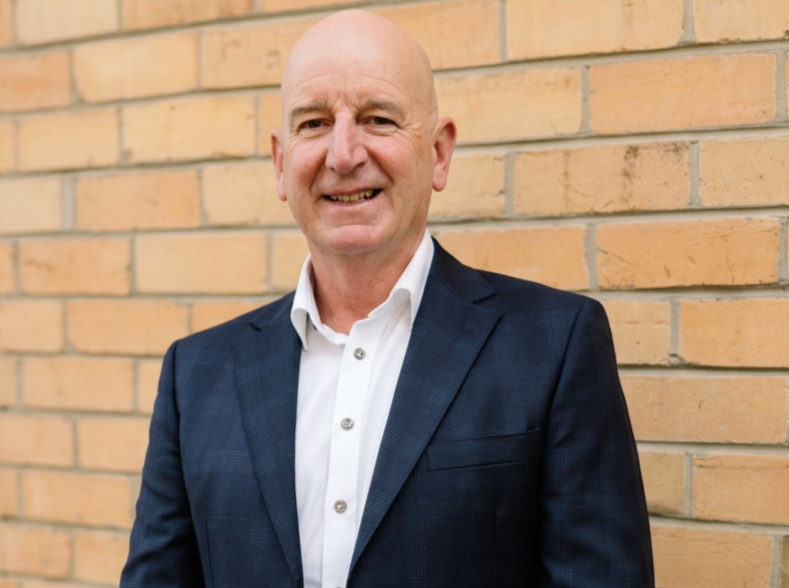 Portrait of a bald man in a dark blazer and white shirt standing against a yellow brick wall.