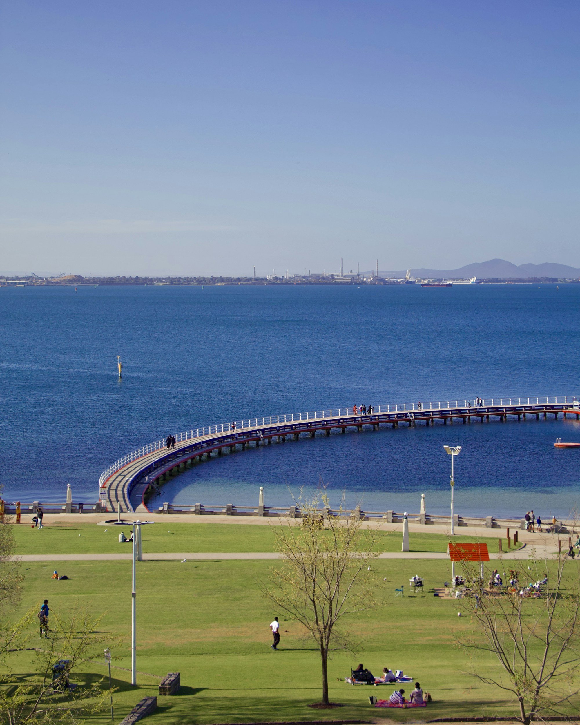 A pier extends into a large body of water with people walking on it. There are grassy areas with people sitting and relaxing, and trees scattered around. In the background, there is an industrial area across the water with buildings and factory smokestacks, and distant mountains.