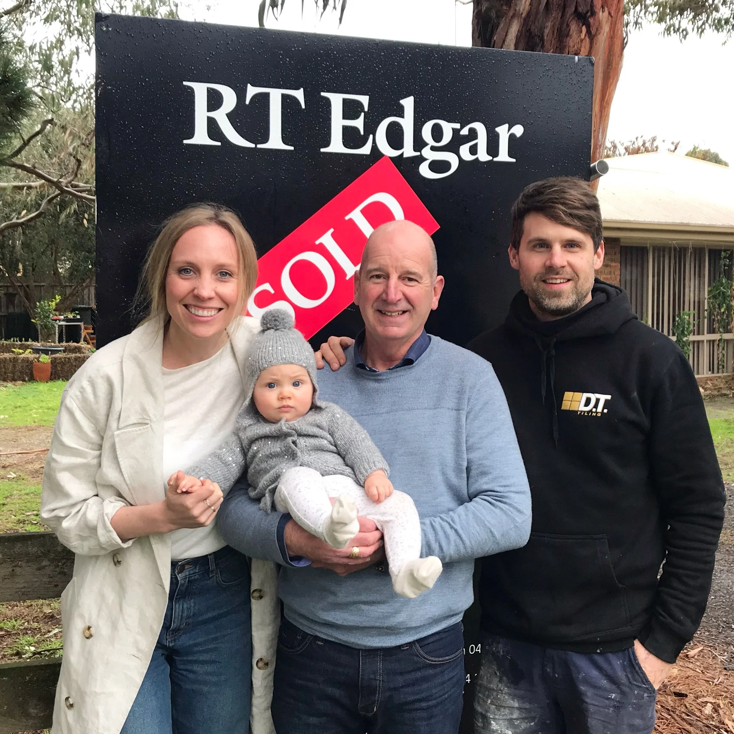 People standing outdoors in front of a black sign with white text that reads 'RT Edgar' and a red 'SOLD' sticker, with trees and a house in the background.