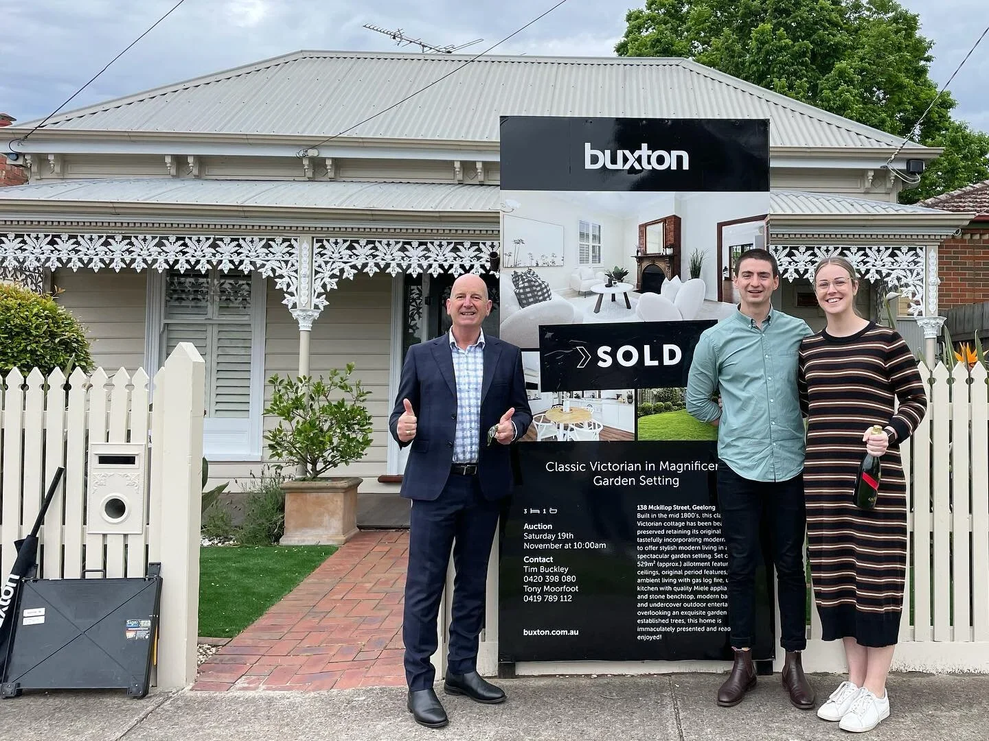 Three people standing in front of a house with a sold sign. The house has Victorian style architecture with decorative white trim, and there is a large sign for Buxton real estate. The man on the left is smiling and giving a thumbs-up, the man in the middle is smiling, and the woman on the right is holding a bottle and smiling.