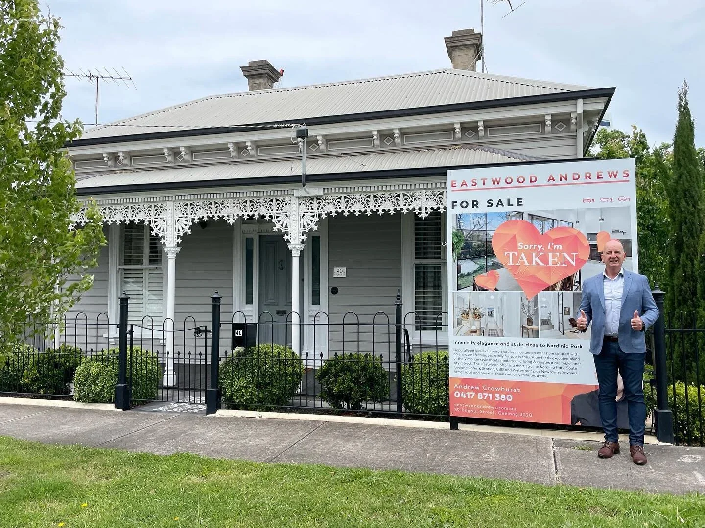 A man in a blue suit giving a double thumbs up standing next to a for-sale sign on a lawn in front of a grey house with ornate white trim and a black fence. The sign reads 'Eastwood Andrews for Sale', with a message about the house and contact details at the bottom.