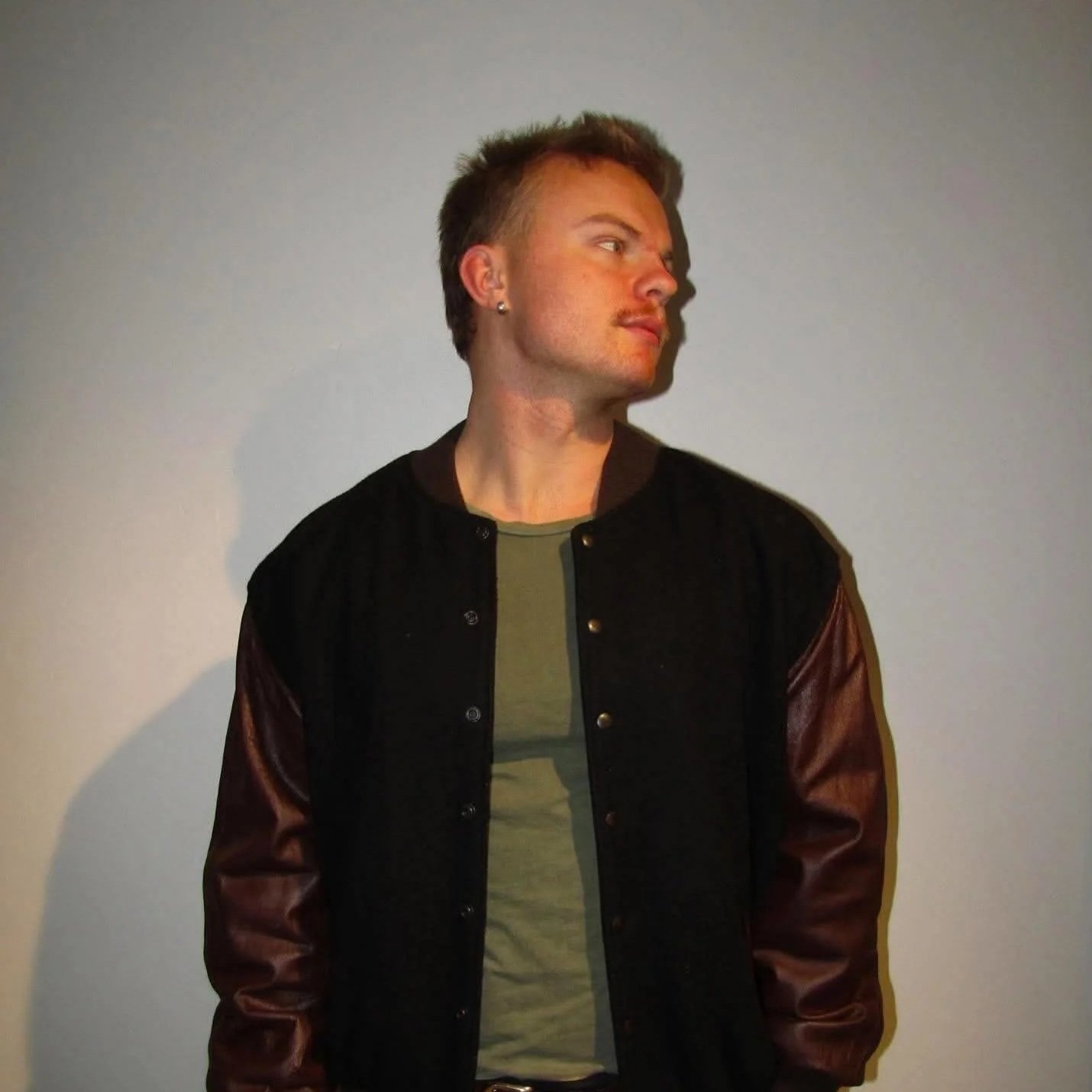 A young man with short brown hair and earrings stands against a plain light gray wall, looking to his left. He is wearing a black jacket with brown leather sleeves over a green t-shirt.