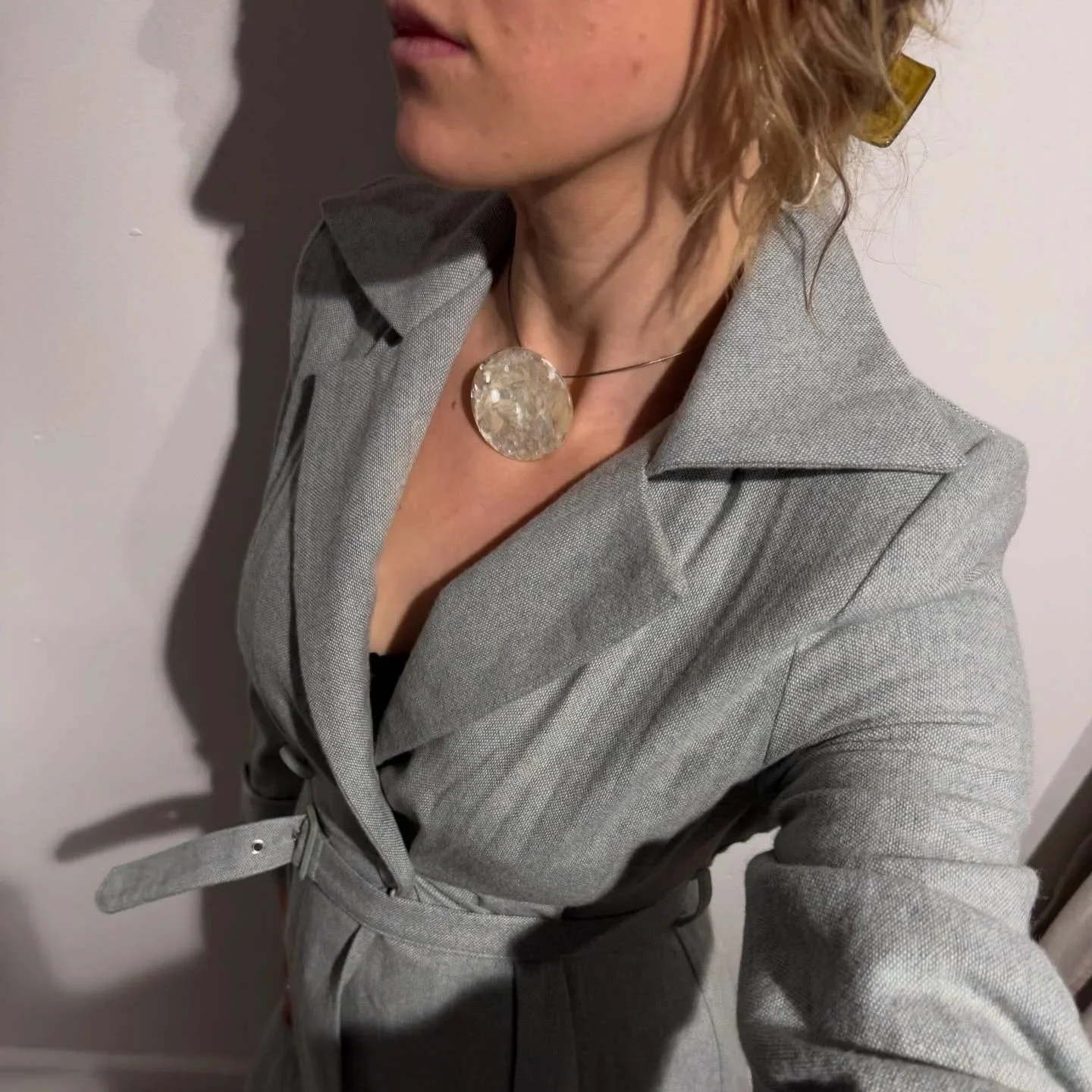A woman wearing a light gray coat with a belt, a black top, and a large circular pendant necklace. Her wavy, light brown hair is partially pinned back with a hair clip. The photo shows her from her shoulders up, turned slightly away from the camera, with a plain light-colored wall in the background.