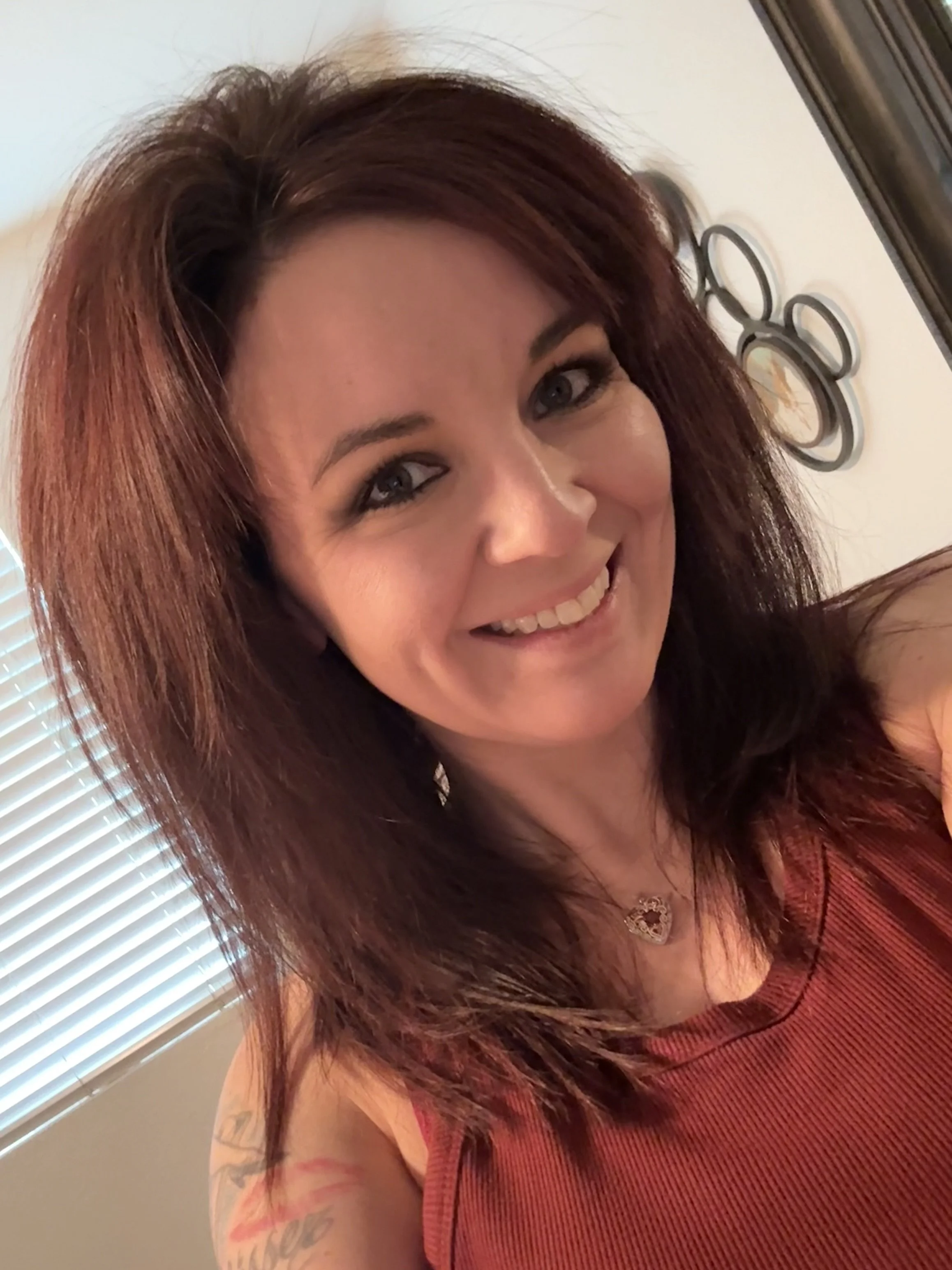 A woman with long reddish-brown hair smiling, wearing a sleeveless orange top, with tattoos on her left arm, and a heart-shaped necklace. Synthwave art