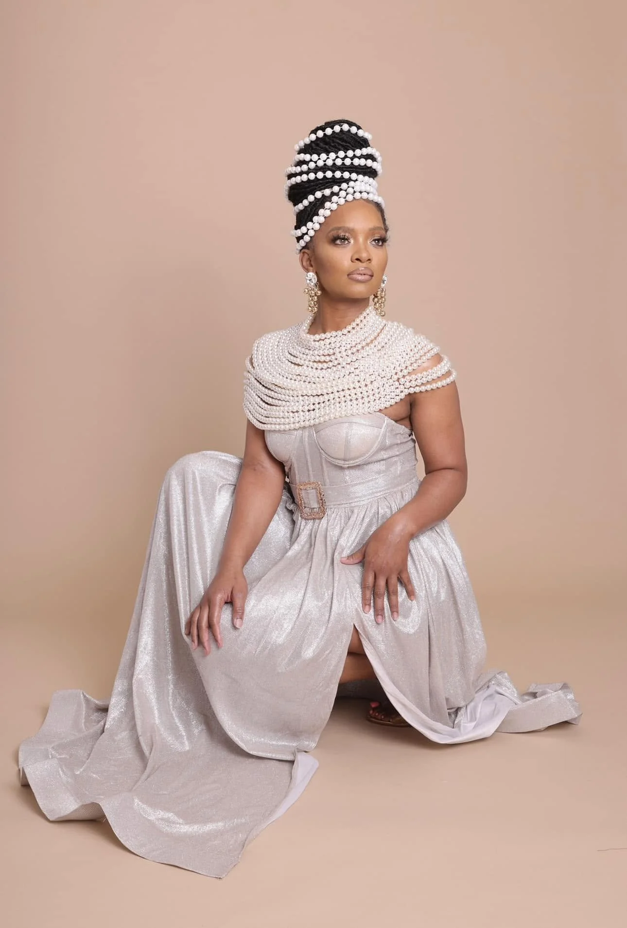 Woman dressed in an elegant gown with layers of pearl necklaces and large earrings, sitting on the floor with a professional studio background.