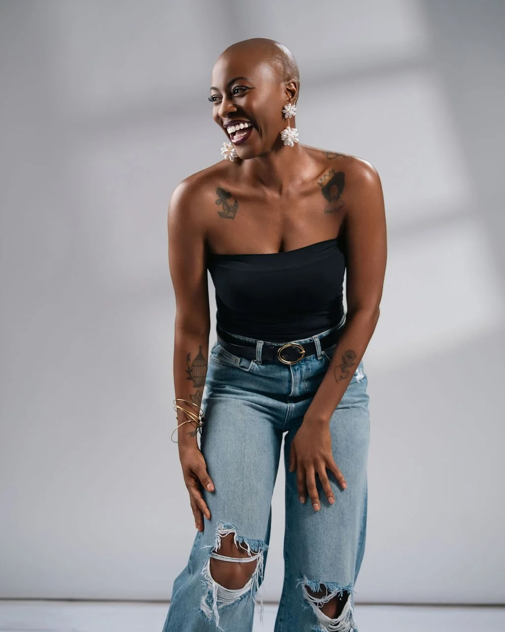 A smiling woman with a shaved head wearing a black strapless top, ripped jeans, and big floral earrings, standing against a neutral background.