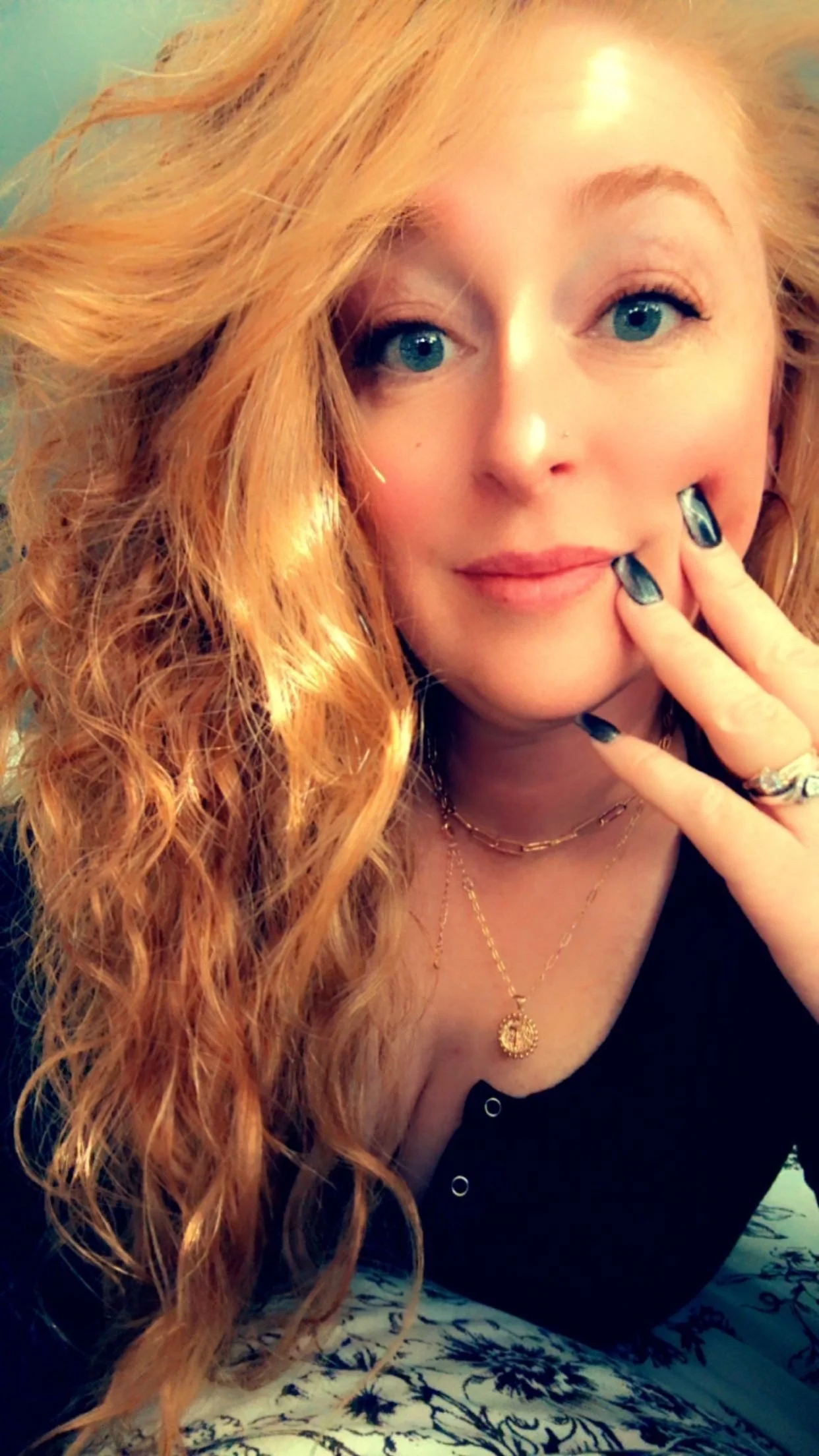 Close-up of a woman with long, curly red hair, blue eyes, and fair skin, smiling softly while touching her lips with her fingers. She is wearing necklaces and rings.