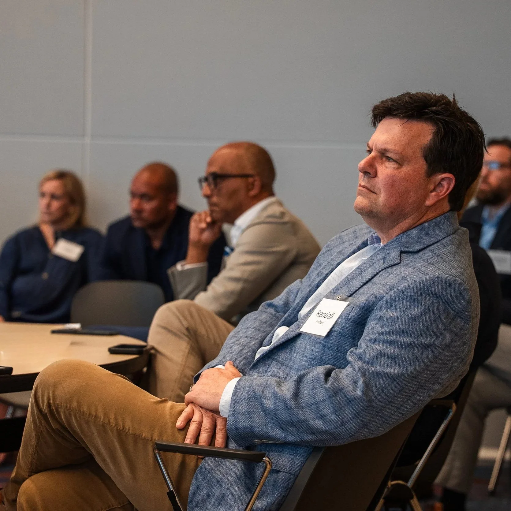 A group of four people sitting in a conference room, paying attention at a presentation. The man in the foreground is wearing a gray blazer and tan pants, with a name tag that says "Randall." Others in the background are women and men dressed in business casual attire, seated against gray walls.