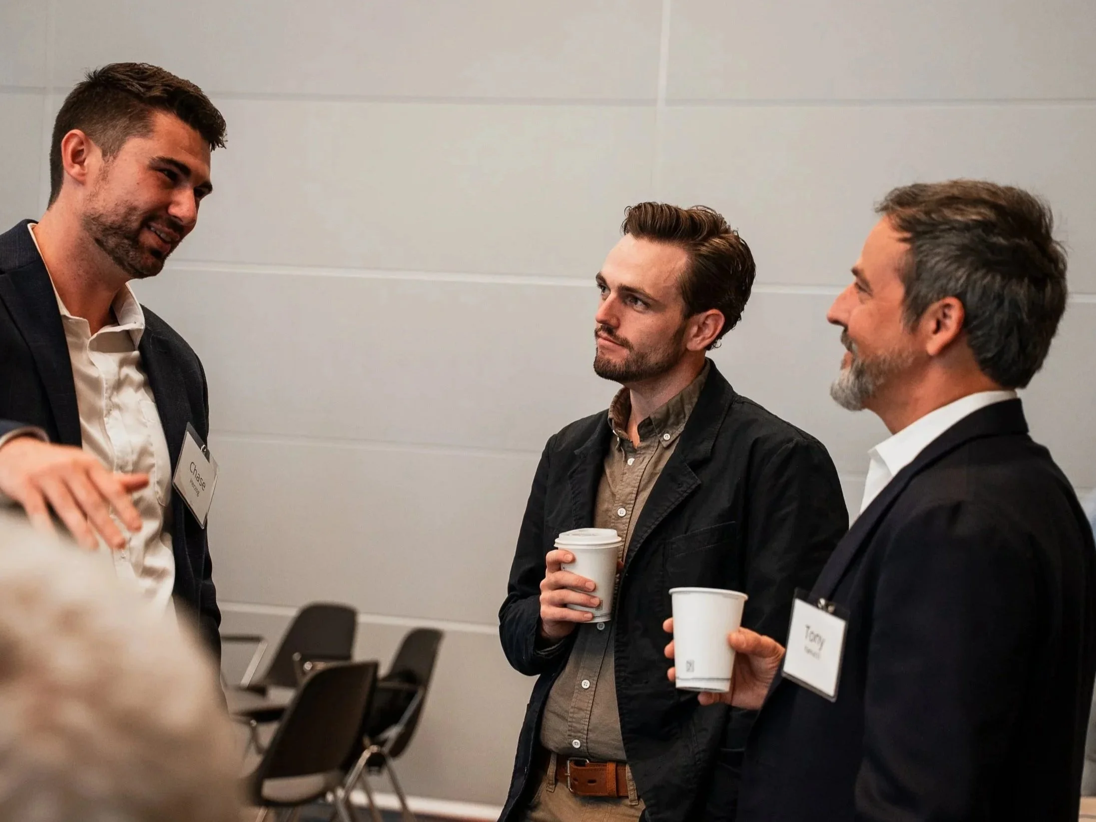 Three men standing in a conference room, engaged in conversation, each holding coffee cups, with name tags visible on their chests.