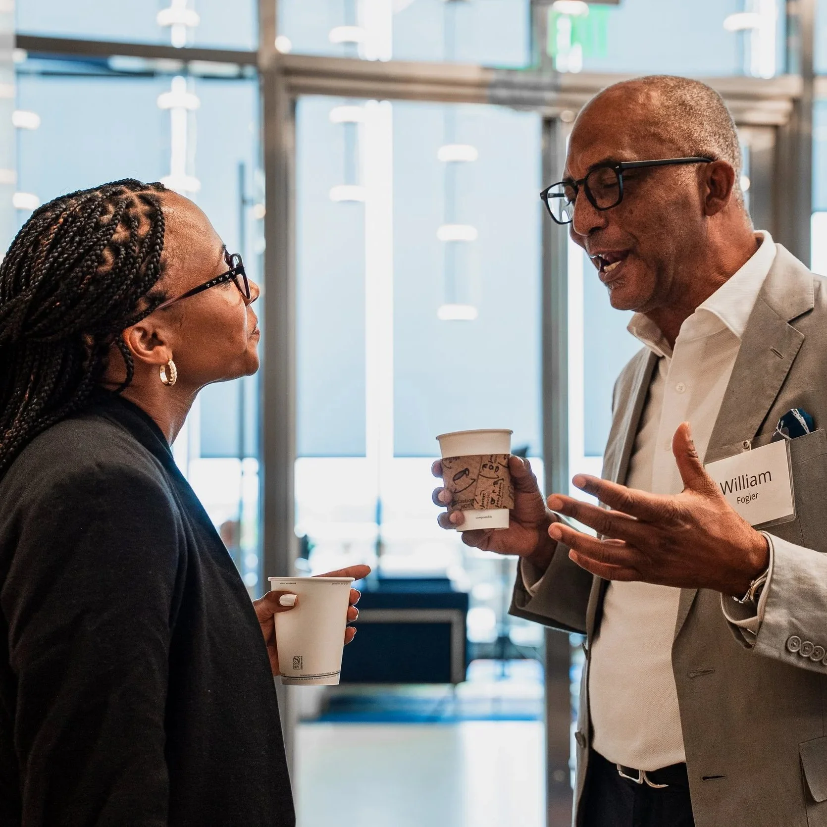 Two professionally dressed people in conversation at an indoor event, both holding coffee cups, with a background of bright windows and modern architecture.