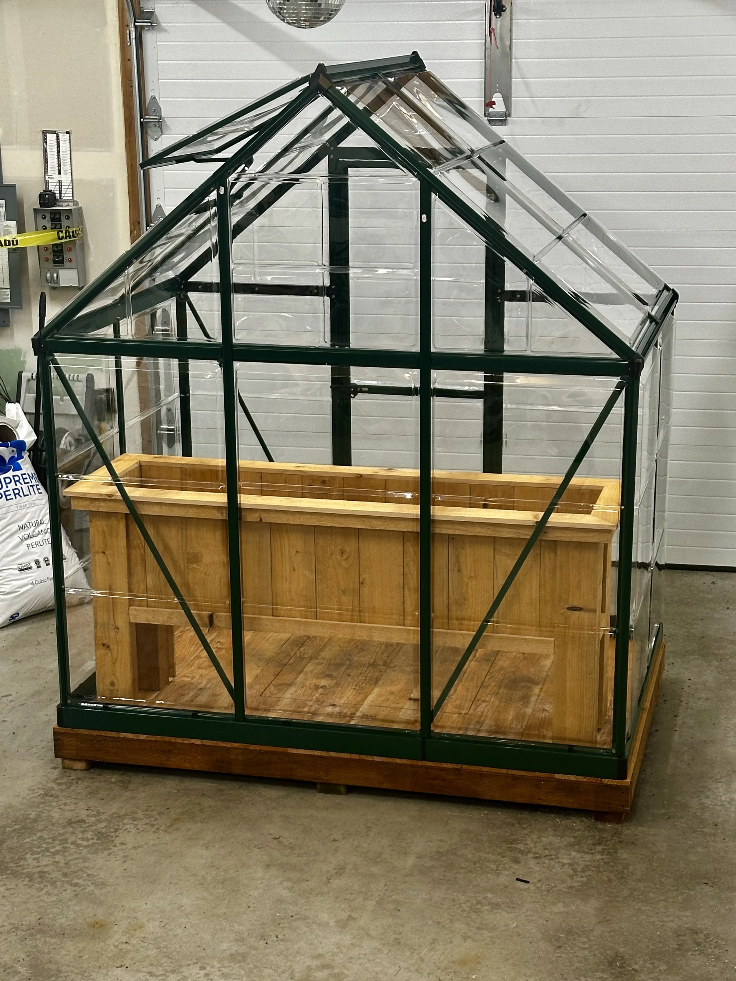 A small greenhouse with a black metal frame, clear glass panels, and a wooden base, situated indoors against a white paneled wall.