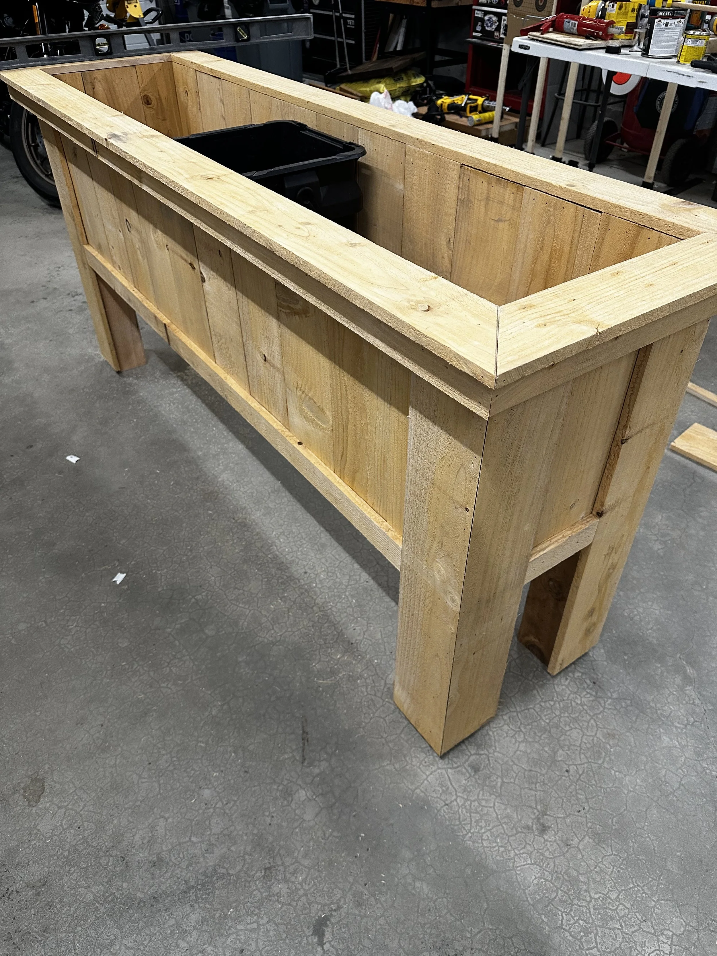 A wooden workbench with a rectangular frame, situated in a workshop or garage. There are tools and supplies on shelves in the background.