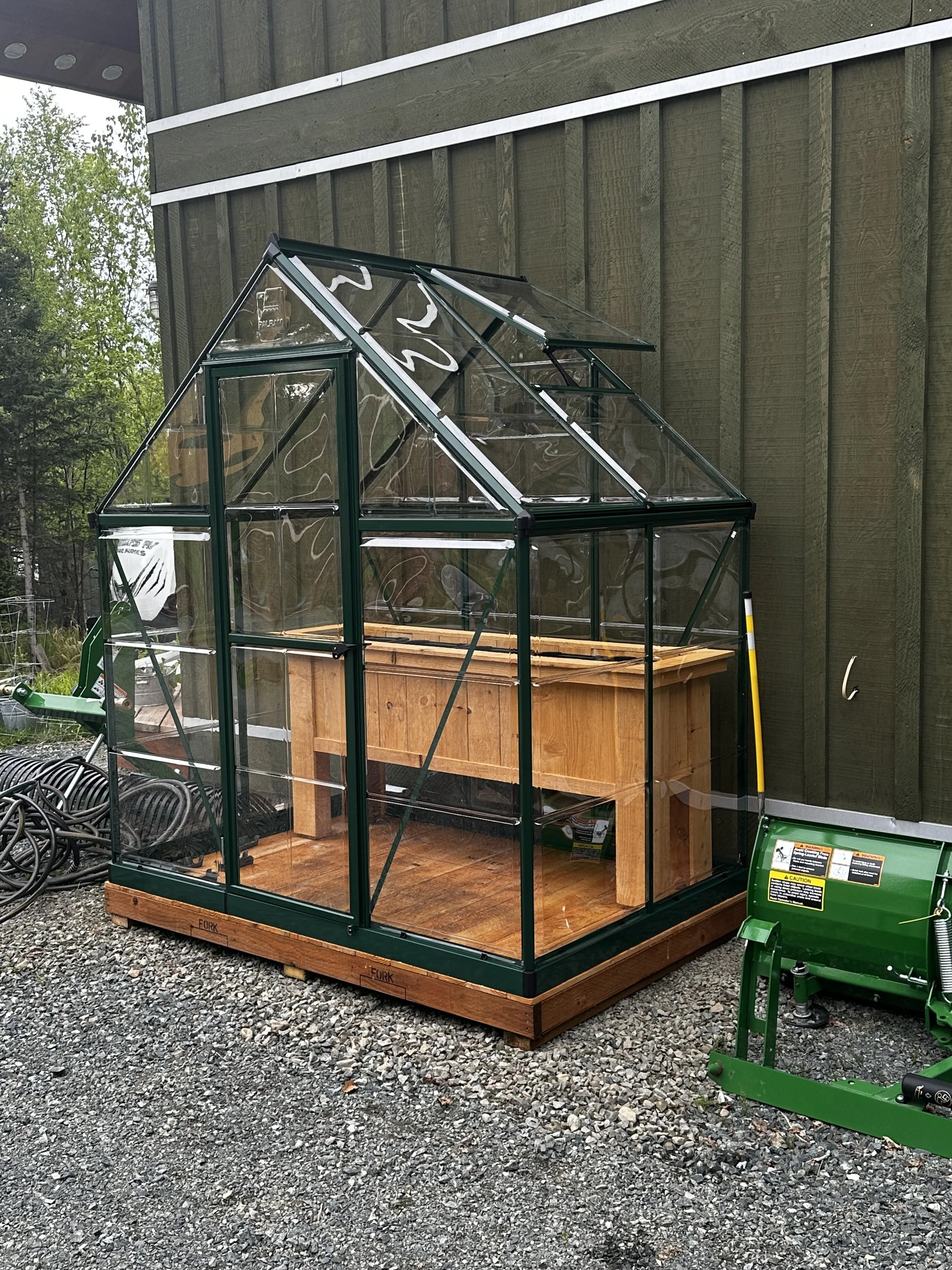 A small greenhouse with a metal frame and clear glass panels, placed on a wooden platform outdoors near a green wall and gravel ground.