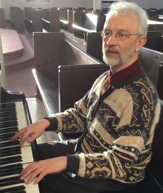 A man with white hair and glasses playing a piano in a church or hall, wearing a patterned sweater.